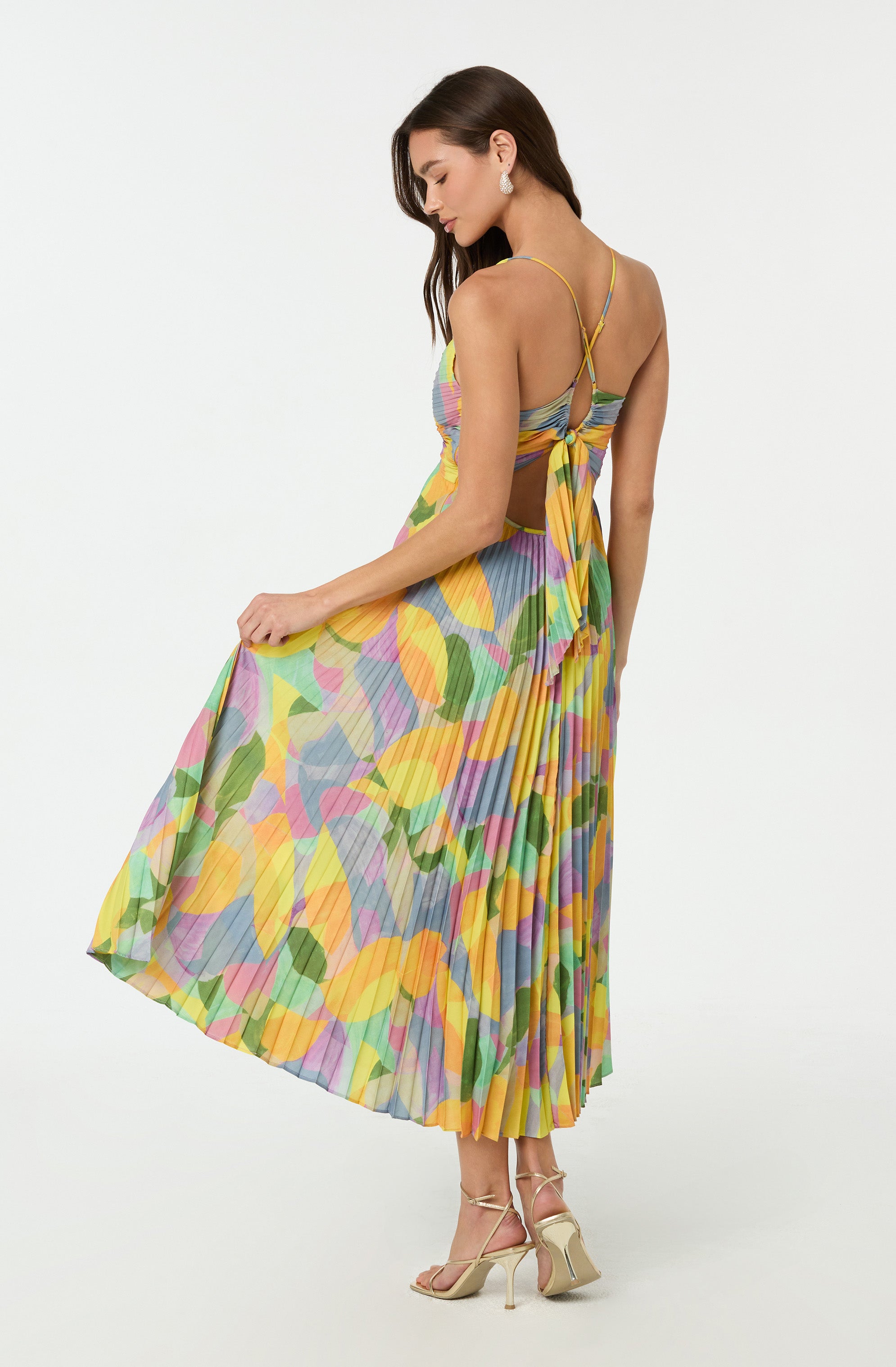 A woman is wearing a colorful, sleeveless, pleated dress with a botanical pattern featuring yellow, pink, green, blue, and purple hues. The dress has thin spaghetti straps that crisscross at the back and a large bow detail just below the shoulder blades. She is paired with beige high-heeled sandals.
