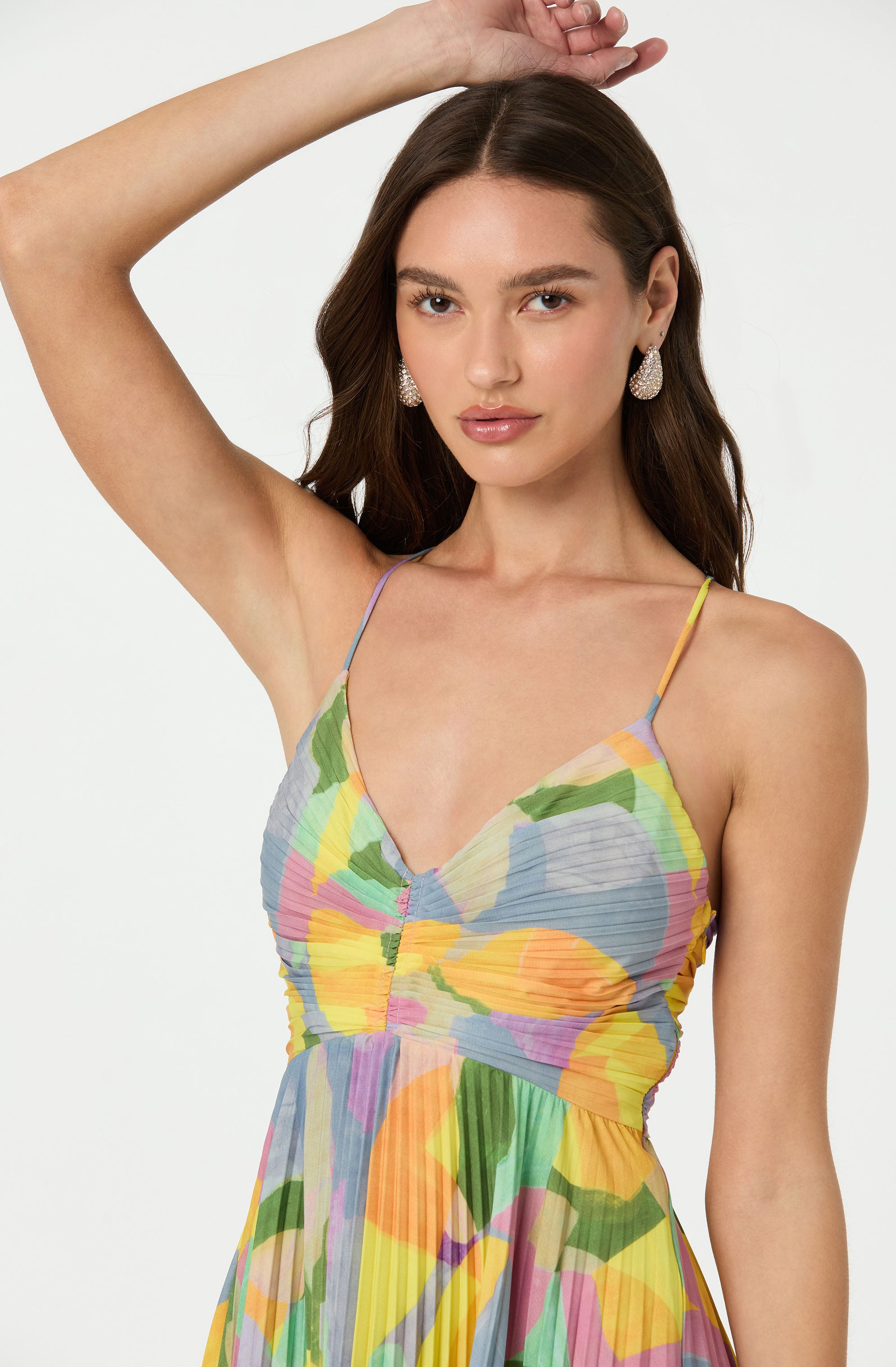 A woman is wearing a colorful, floral-patterned dress with thin spaghetti straps. The dress features a pleated fabric with shades of yellow, green, pink, purple, and orange, creating a vibrant and lively appearance.