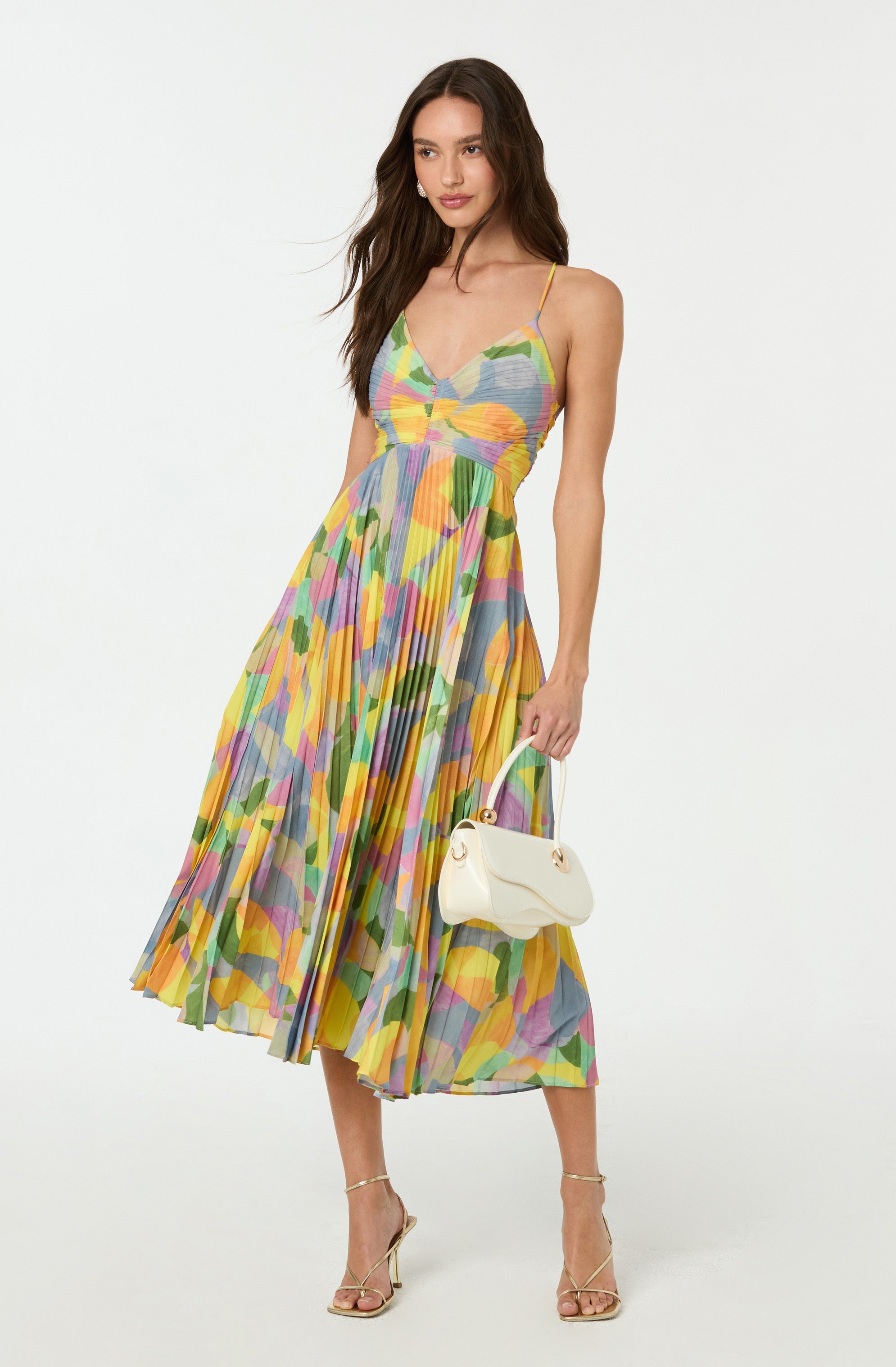 A woman is wearing a colorful, pleated, sleeveless dress with thin spaghetti straps. The dress features an abstract, multicolored pattern with shades of yellow, green, pink, purple, and blue. She is also holding a small, white handbag with a curved design and gold accents, and is wearing beige high-heeled sandals with thin straps.