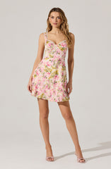 The dress is a sleeveless, above-the-knee style with thin straps adorned with small ruffles. It features a fitted bust with a sweetheart neckline and an all-over floral pattern in shades of pink, green, and white. The fabric appears lightweight and flowy. The model is wearing light pink open-toe high heel shoes.