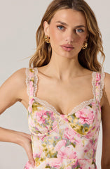 A woman is wearing a sleeveless, floral dress with pink, yellow, and green colors. The dress features lace trim along the neckline and straps, and has a fitted bodice with a sweetheart neckline.