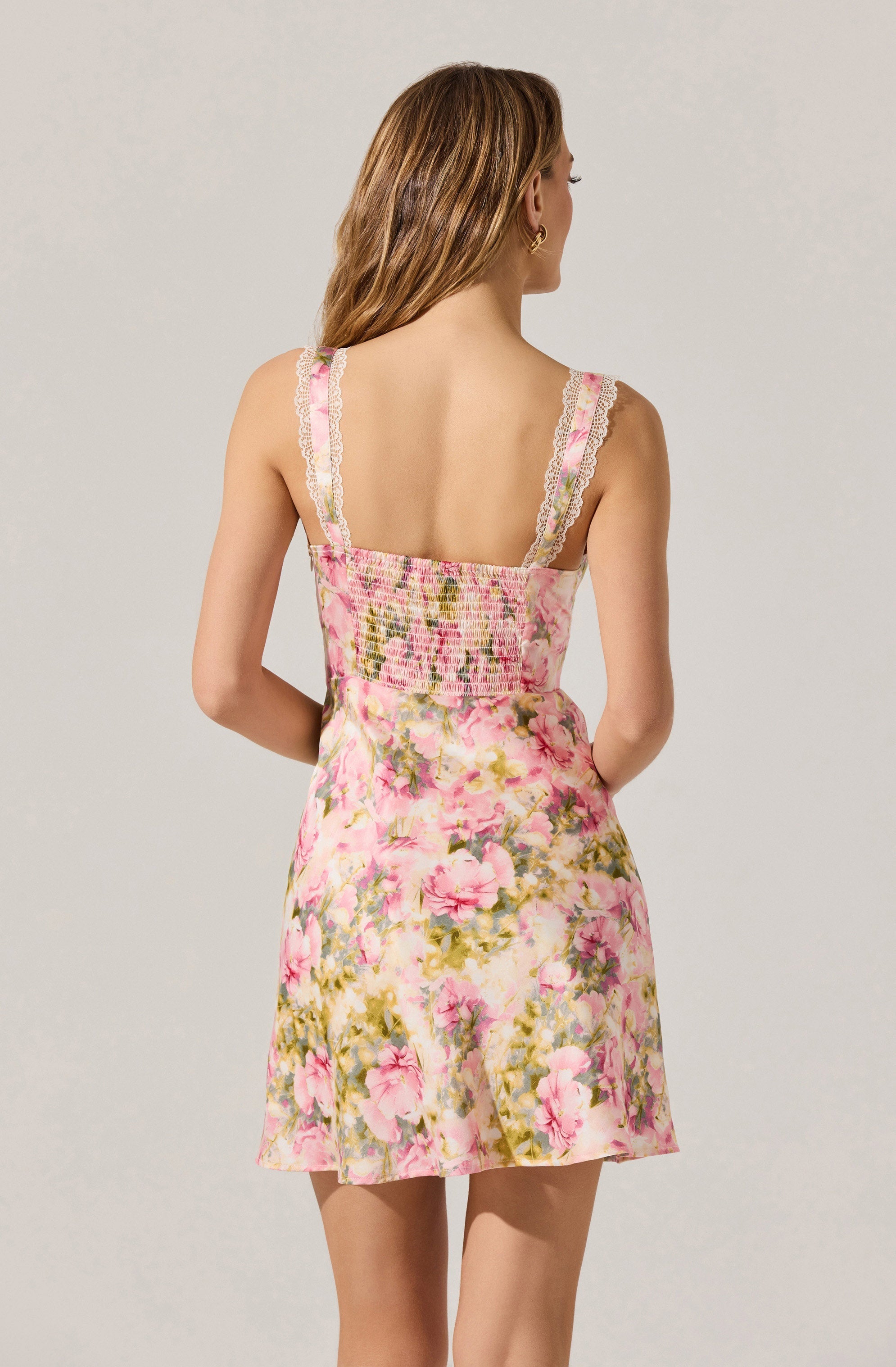 A woman is wearing a sleeveless dress with wide straps decorated with pink and white lace trim. The dress features a smocked bodice and a floral pattern with pink, green, and cream colors on a light background. The skirt of the dress is flared and short.