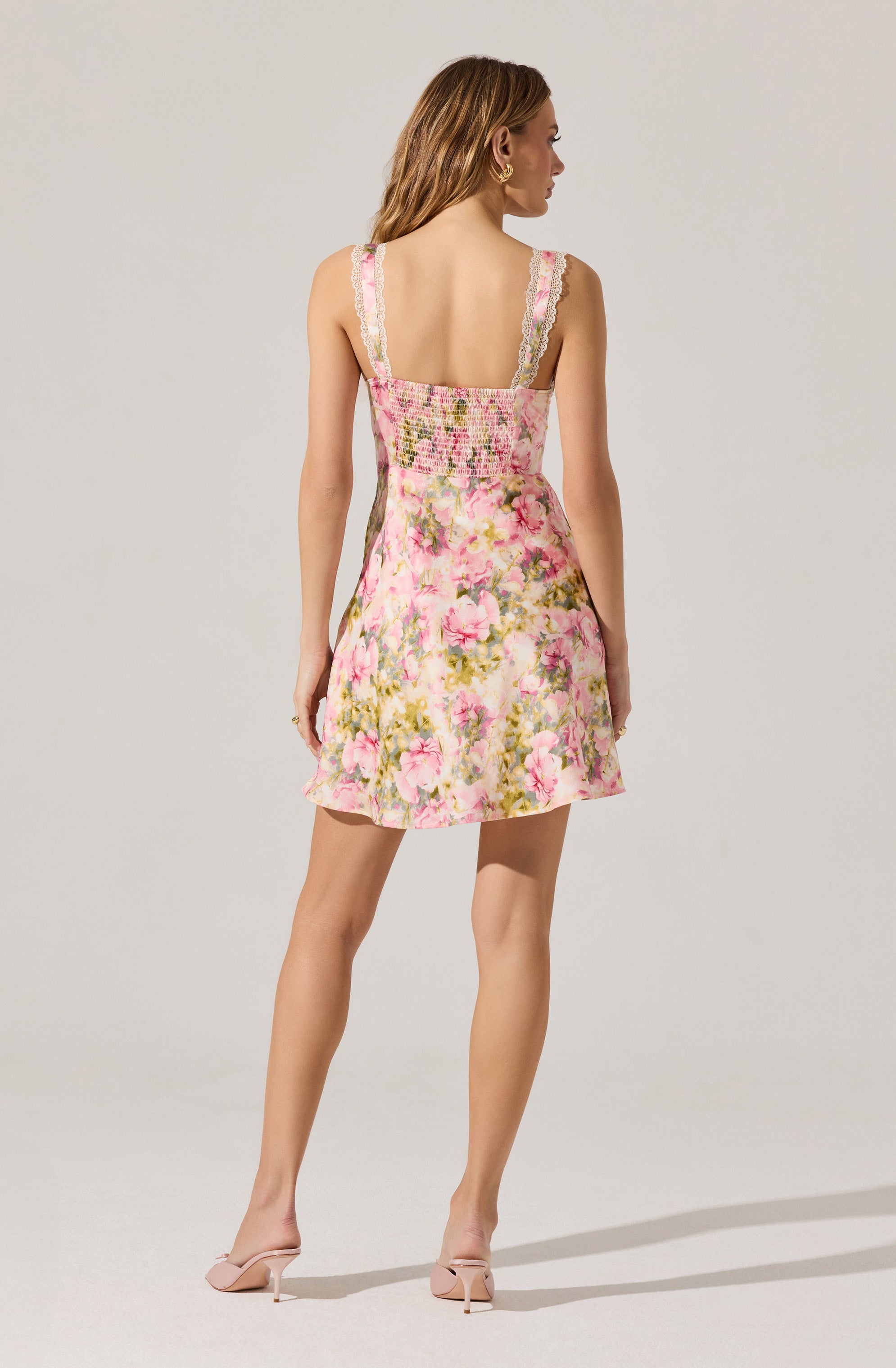 A woman is wearing a sleeveless, ruffled-strap dress with a fitted, shirred bodice and a short, flared skirt. The dress features a floral pattern with pink, green, and white tones.