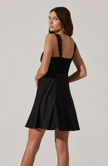 A woman is wearing a black sleeveless dress with wide shoulder straps and lace detailing along the straps. The dress has a fitted bodice and a flared skirt that falls above the knees. The fabric appears smooth and slightly shiny, giving it an elegant look.