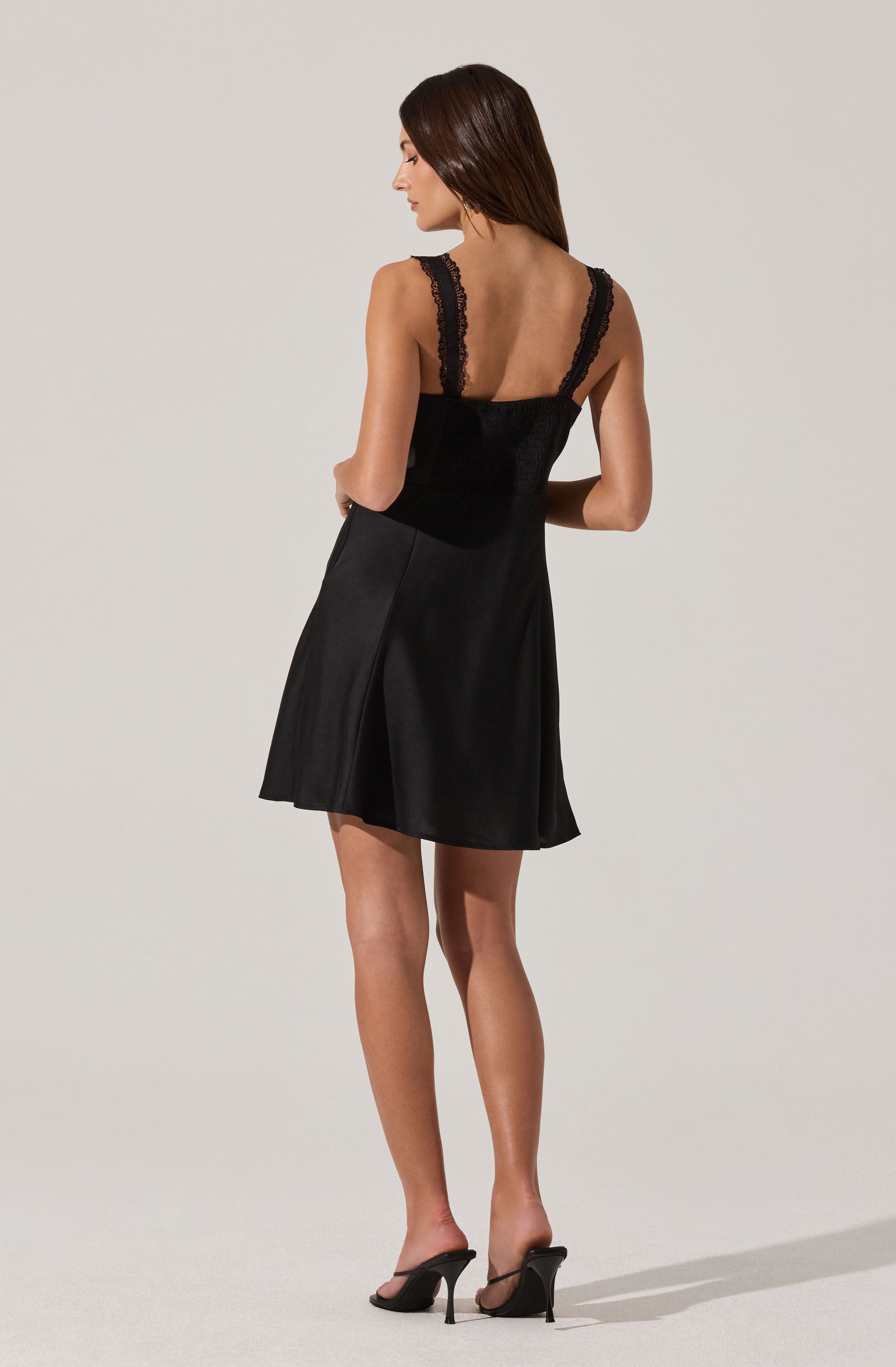 A woman is wearing a black sleeveless dress with lace straps on the shoulders. The dress has a fitted bodice and a flared skirt that falls above the knees. She is also wearing black high-heeled sandals with thin straps.