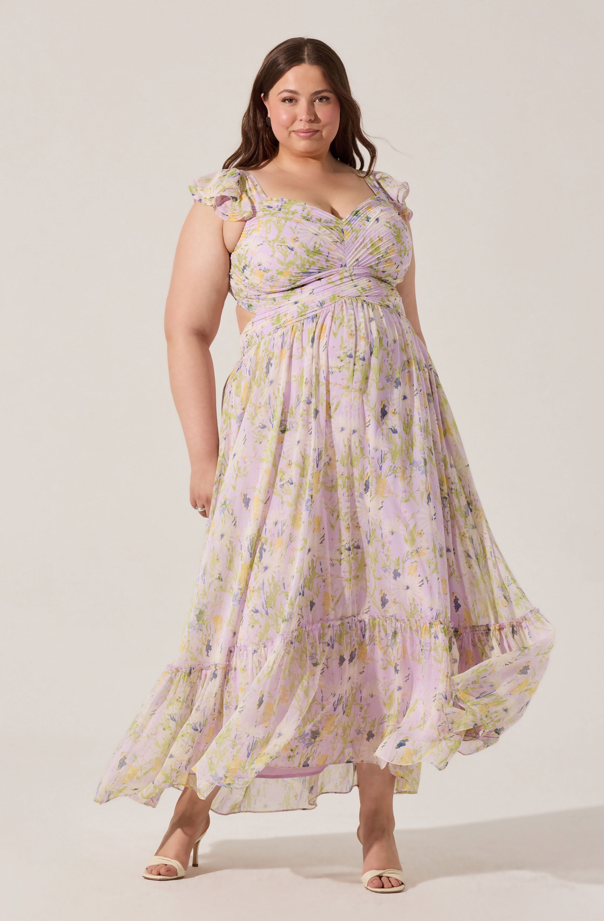 A woman is wearing a long, flowy dress with a pastel floral pattern consisting of light pink, yellow, purple, and green colors. The dress has short, ruffled sleeves and a fitted bodice with gathered fabric at the bust, creating a ruched effect. The skirt is tiered, with a ruffle at the bottom hem, and the fabric appears lightweight and airy. She is also wearing open-toed, beige high heel shoes.