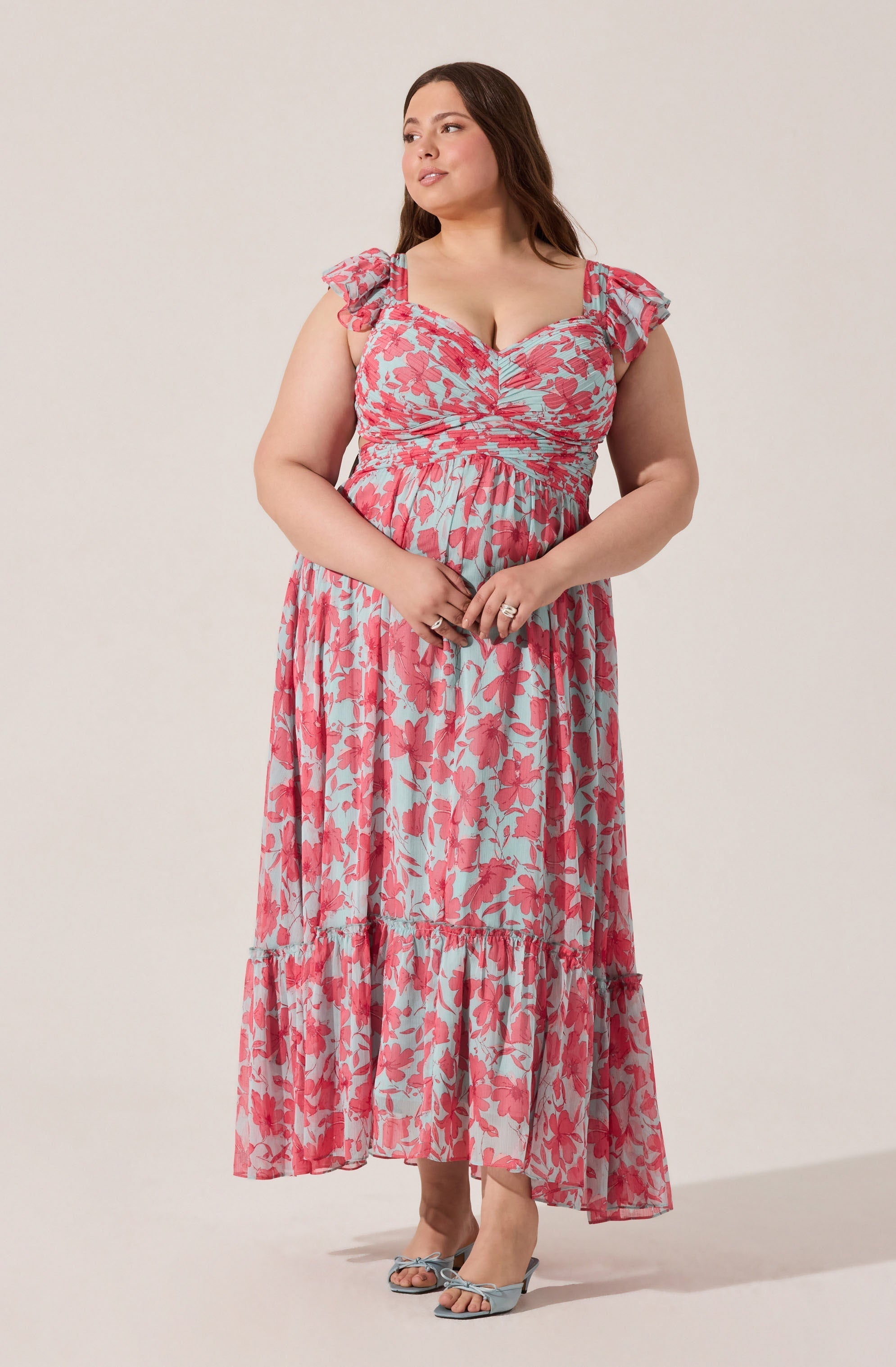 A woman is wearing a long, flowy dress with a pink and light blue floral pattern. The dress has ruffled shoulder straps and a fitted bodice. She is also wearing open-toed high-heeled shoes in a matching light blue color with bows on the front.