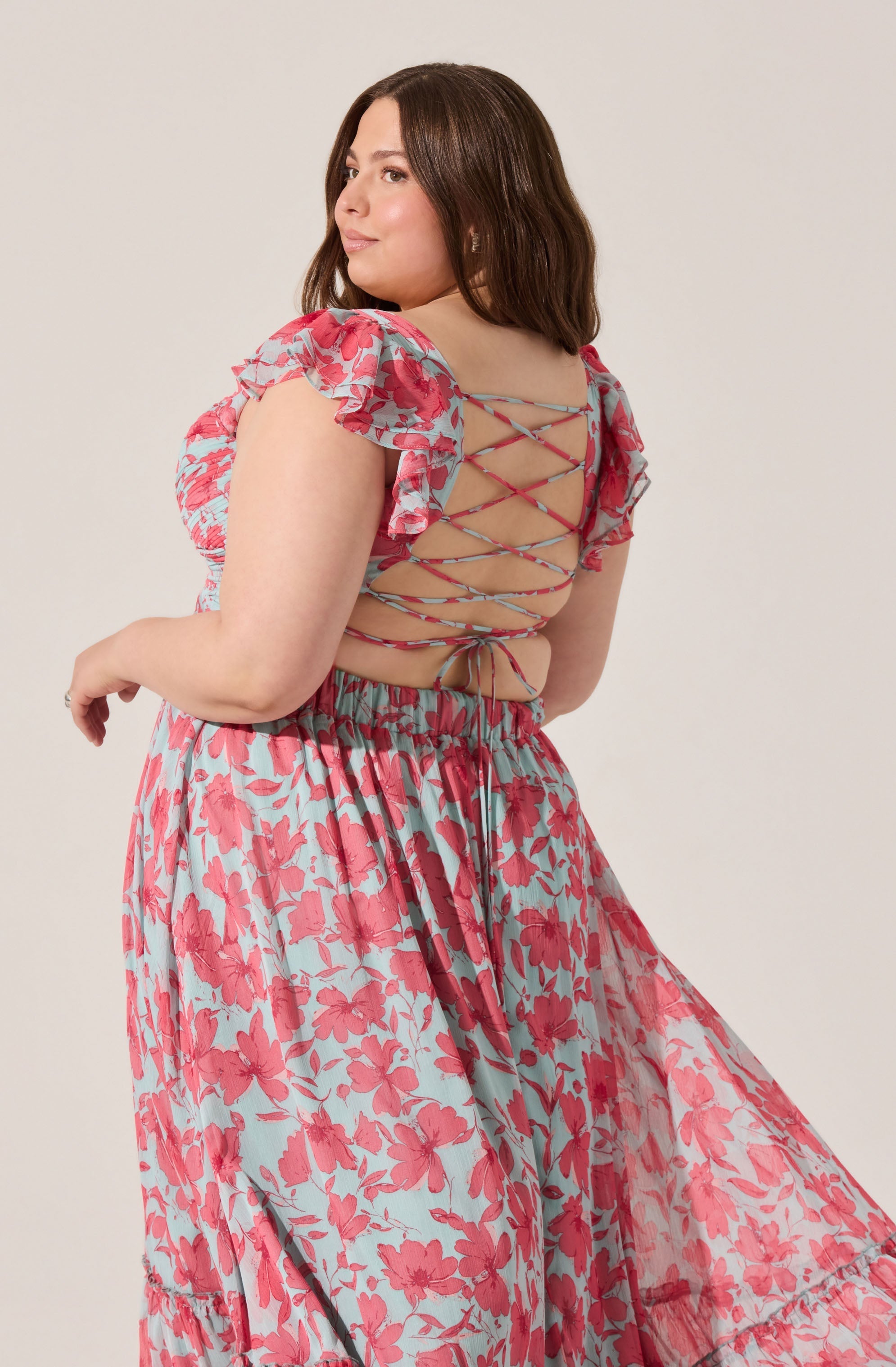 A woman is wearing a light blue dress with a red floral pattern. The dress features ruffled short sleeves and an open back with crisscrossing ties that lace up along the back. The skirt is long and flowing with a gathered waistband.
