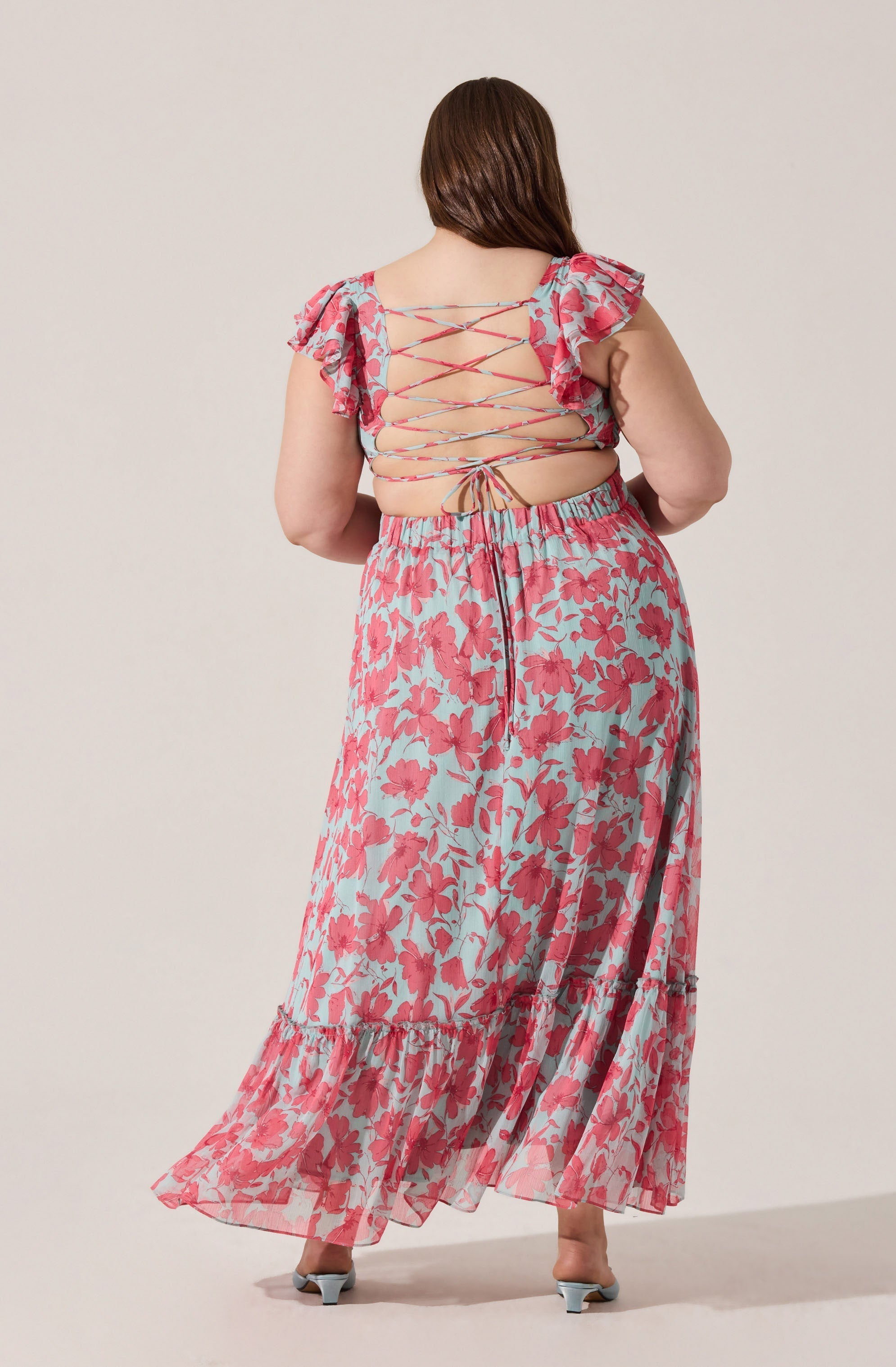 A woman is wearing a two-piece outfit with a floral pattern. The top features a lace-up back with thin crisscrossing ties and ruffled cap sleeves. The matching long skirt has the same floral print and a tiered ruffle hem. She is also wearing open-toed heels.