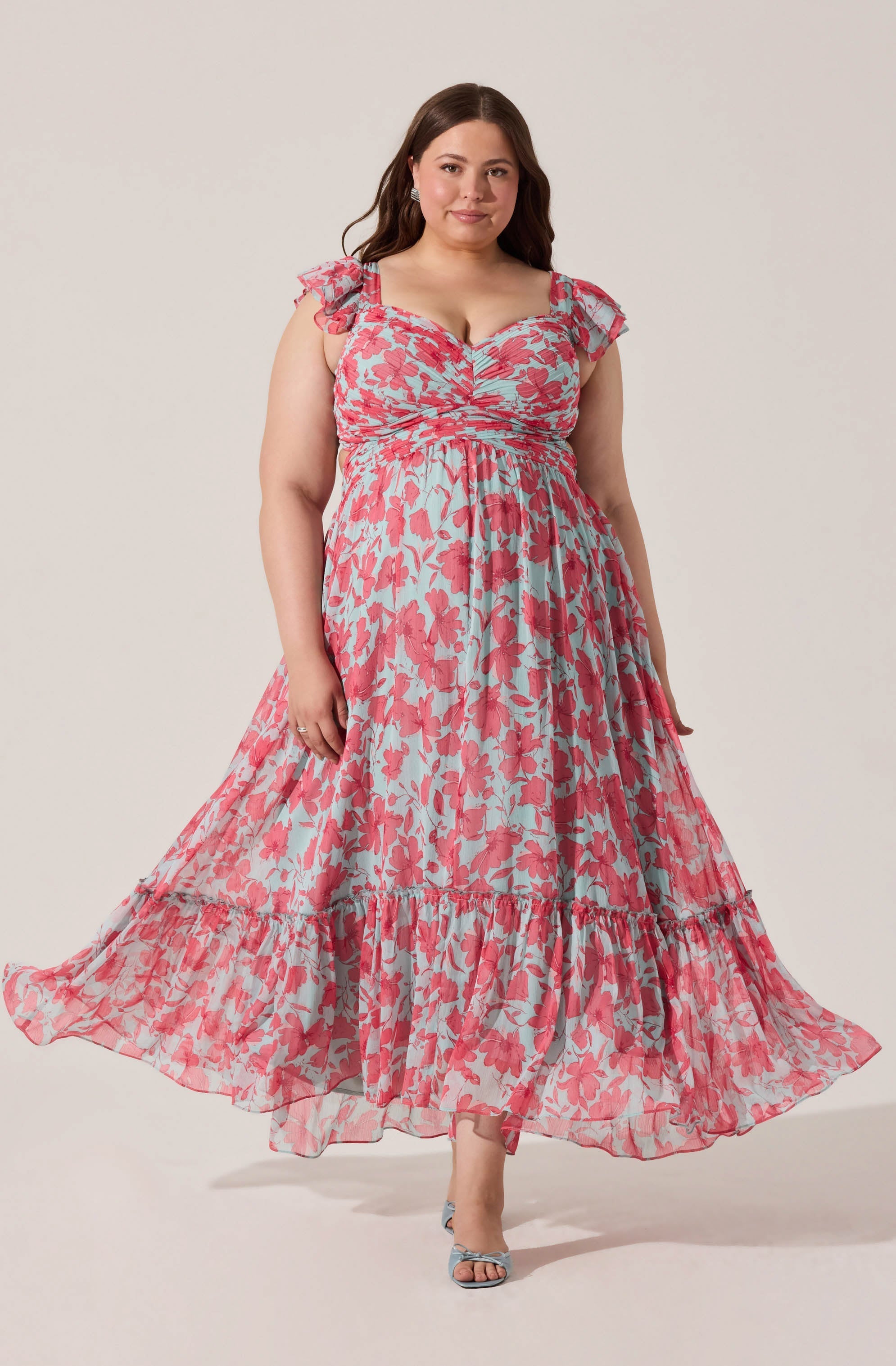A woman is wearing a long, flowing dress with a light blue background and a pink floral pattern. The dress features ruffled shoulder straps, a ruched bodice, and a tiered hemline. She is also wearing open-toed high heels with a bow detail.