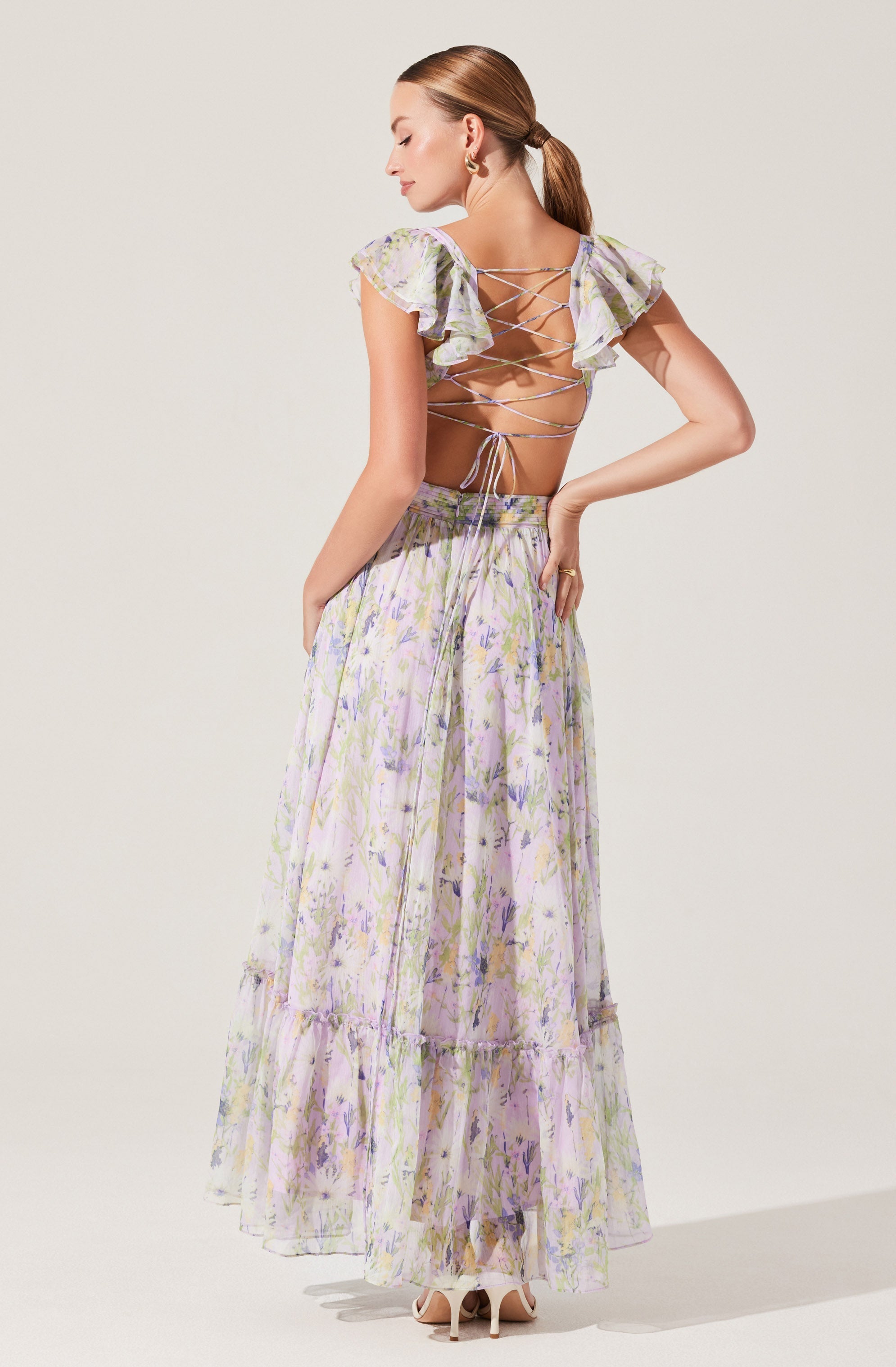 A woman wearing a two-piece outfit with a pastel floral pattern. The top features ruffled short sleeves and a back laced-up design with crisscrossing ties. The long, flowing skirt also displays a matching floral pattern and has a tiered ruffle detail near the hem. She is wearing high-heeled shoes and has pulled her hair back into a ponytail.