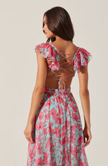 A young woman is wearing a matching floral outfit with a sleeveless top and a long skirt. The top features ruffled, semi-transparent sleeves and a crisscross lace-up back detail. The high-waisted skirt has an elastic waistband and a pink floral pattern on a light blue background.