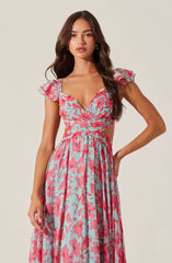 A woman is wearing a long, flowing dress with a floral pattern in pink and light blue. The dress features ruffled short sleeves, a ruched bodice with a sweetheart neckline, and a waistline that accentuates her figure.