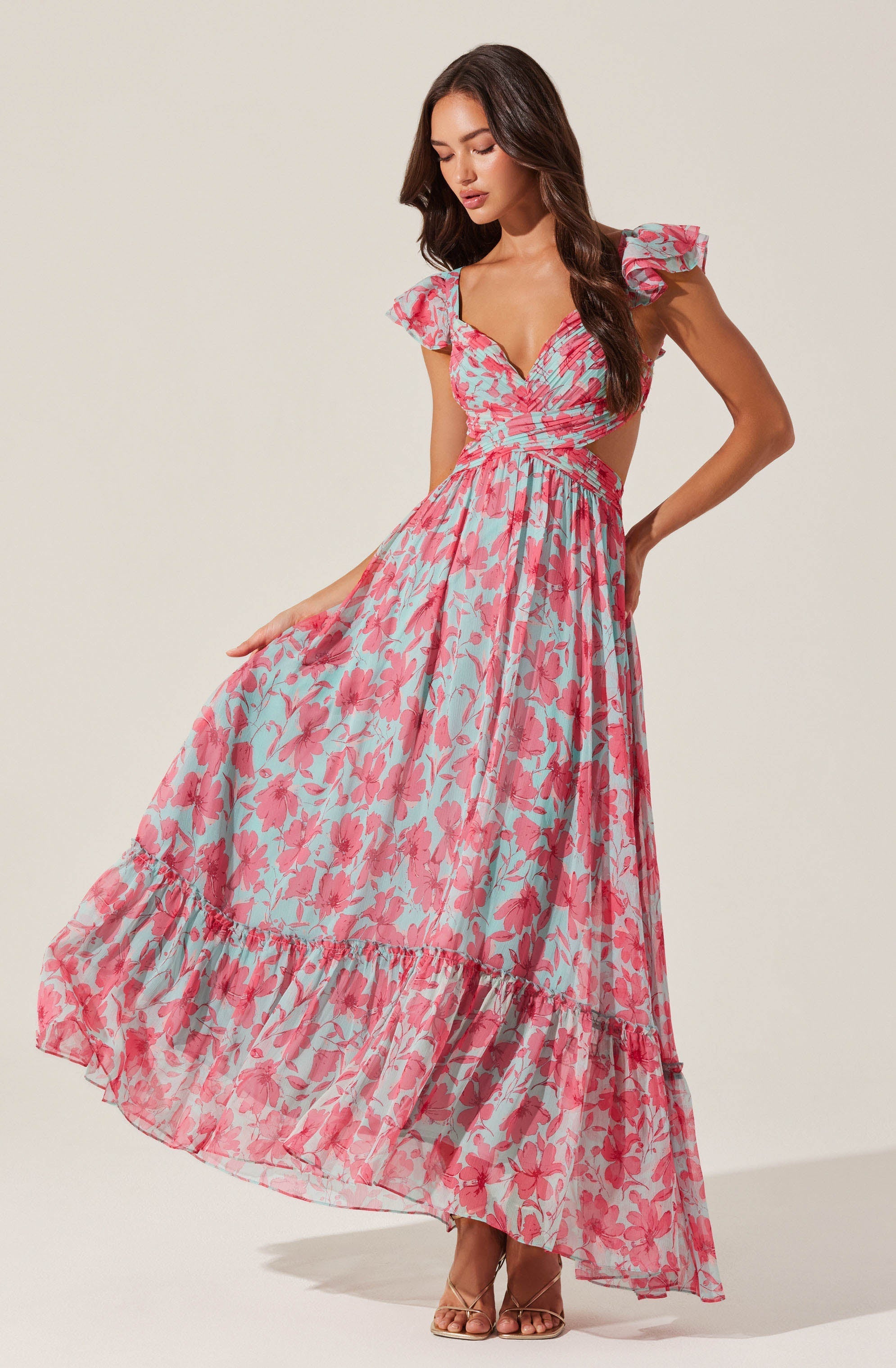 A woman is wearing a long, flowing dress with a pink and light blue floral pattern. The dress features ruffled cap sleeves, a fitted bodice with a V-neckline, and cut-out sides at the waist. The skirt has a tiered design with ruffles at the hem, creating a breezy, summery look.