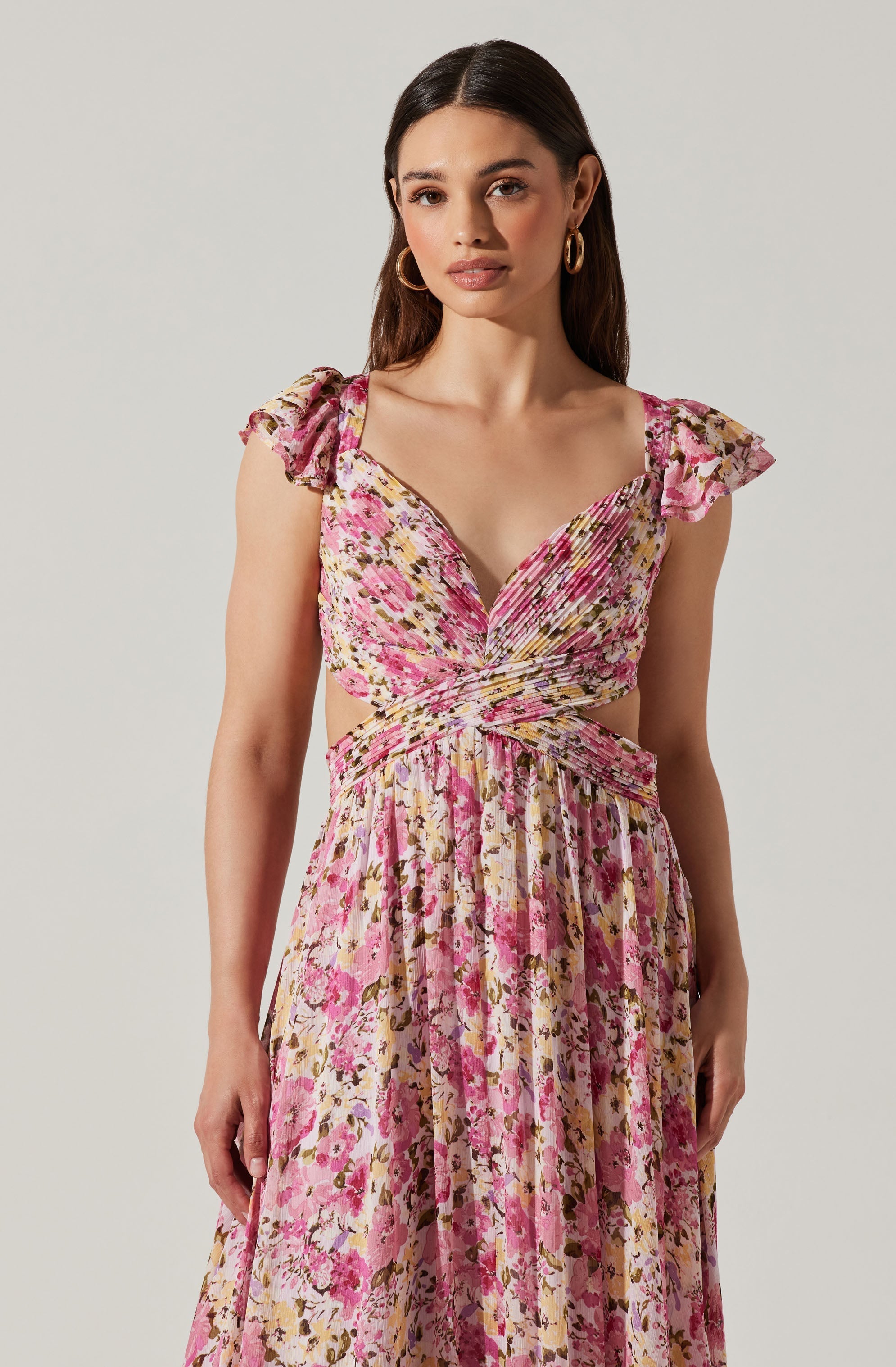 A woman is wearing a long, flowing dress with a floral pattern in shades of pink, yellow, and brown. The dress has ruffled, flutter sleeves and a neckline that crosses at the chest, creating a gathered effect at the waist. The fabric features a mix of small and large floral designs, and the dress has a pleated skirt that extends downwards.