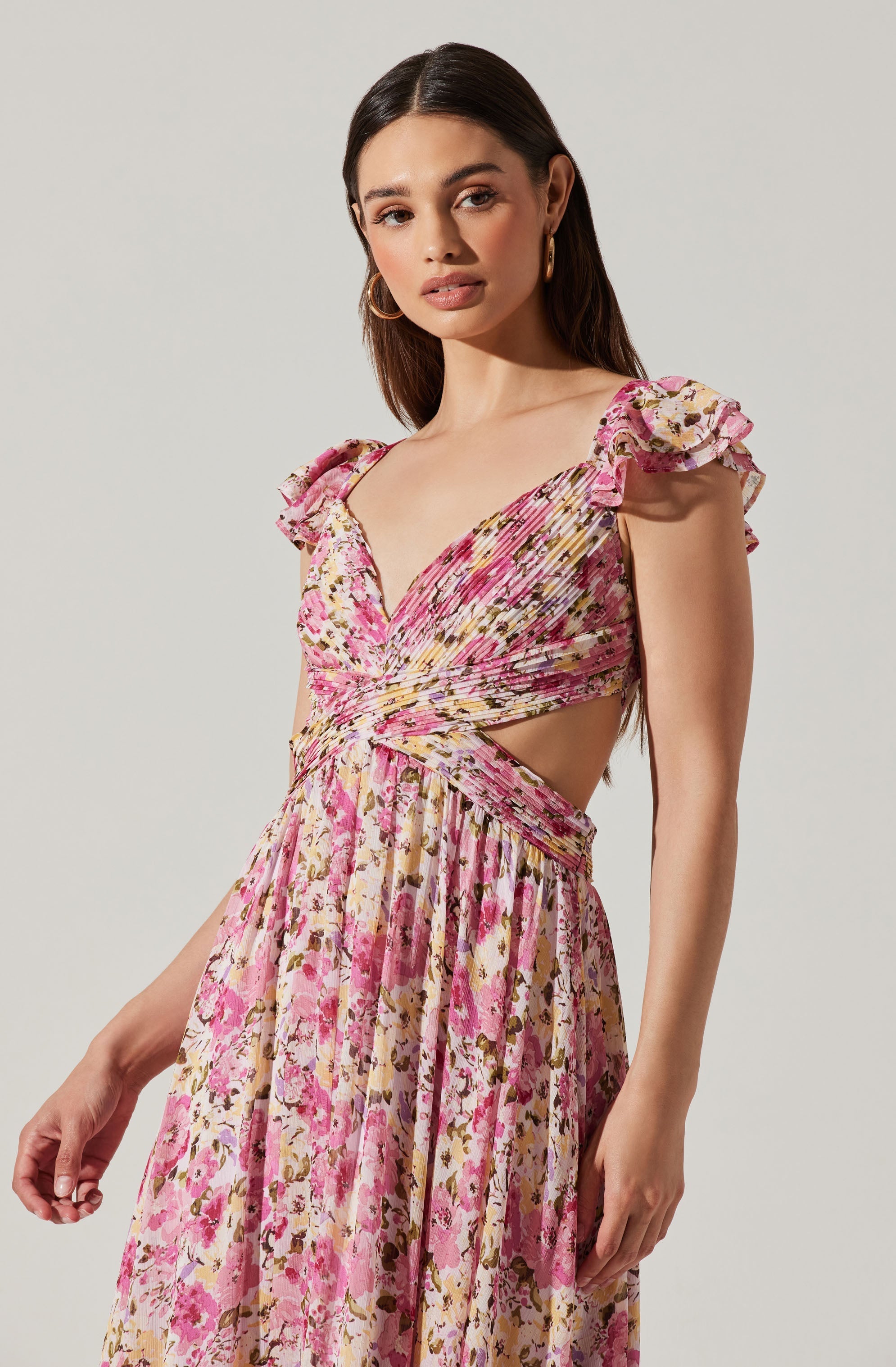 A woman is wearing a pink floral dress with ruffled cap sleeves and a cutout at the waist. The dress has a pleated, flowing skirt with a multicolored floral pattern.