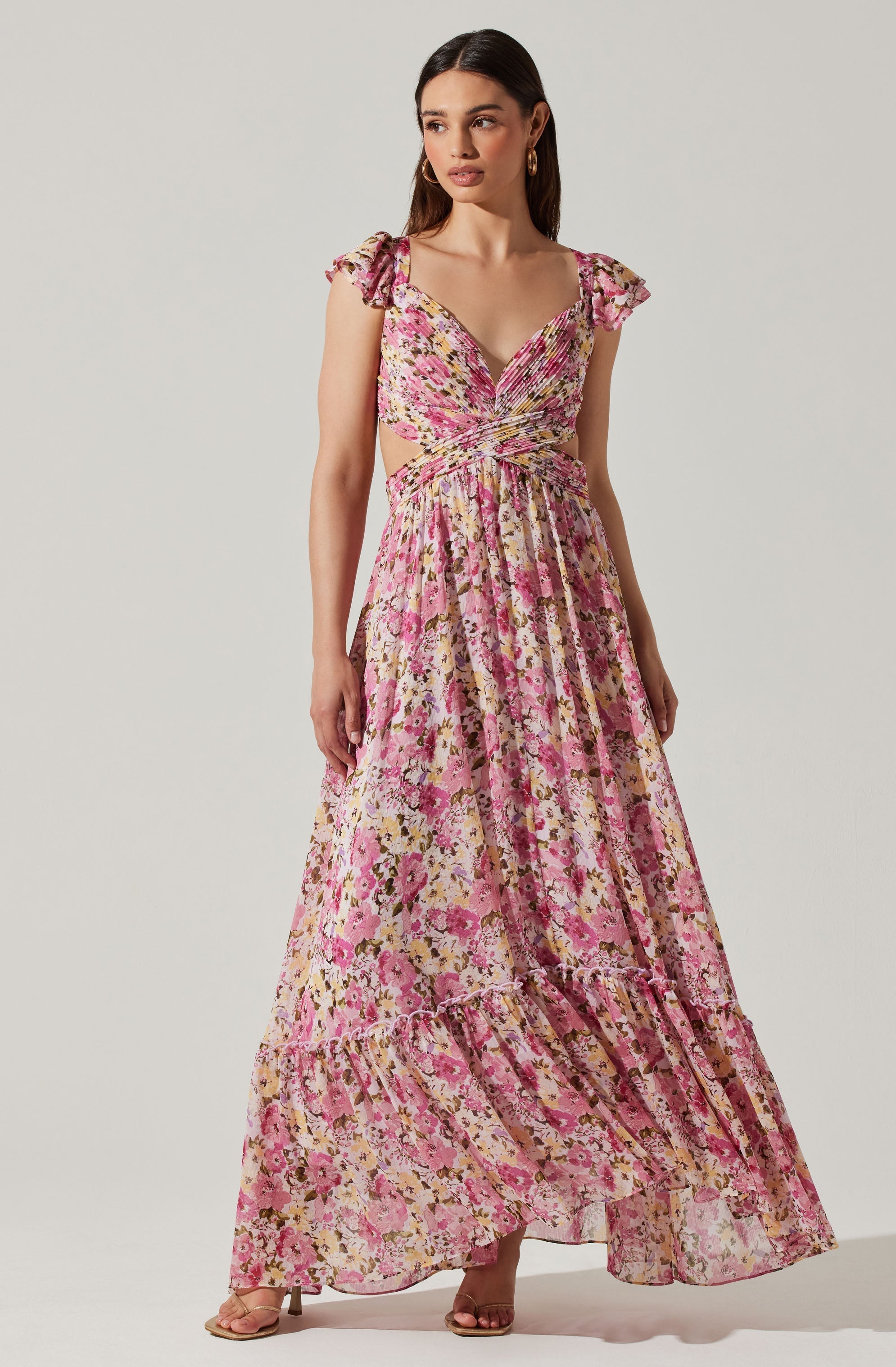 A woman is wearing a long, flowing floral dress with shades of pink, beige, and hints of yellow. The dress features short, ruffled sleeves and a ruched bodice with cutouts at the waist.