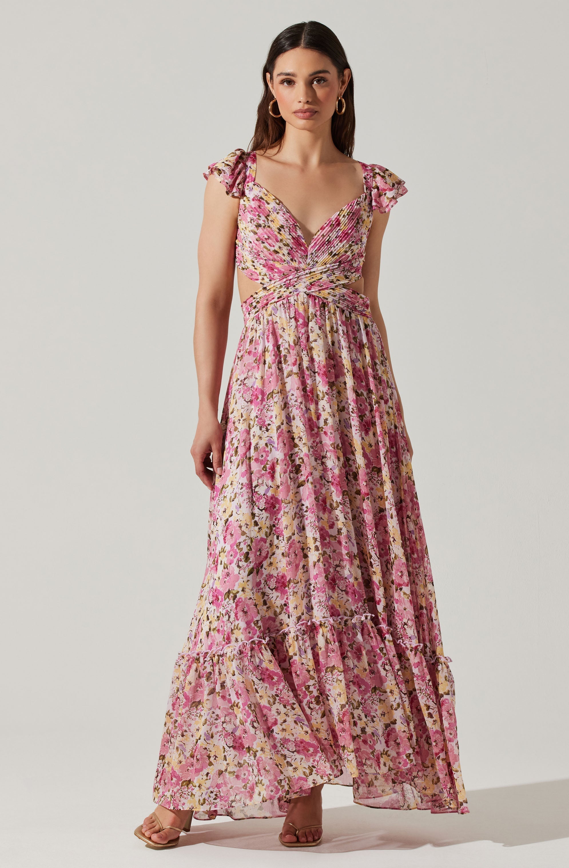 A woman is wearing a long, flowing dress with a pink, yellow, and brown floral pattern. The dress features a sweetheart neckline with ruched detailing at the bust, ruffled cap sleeves, and cut-out sides at the waist. The skirt is gathered at the waist and has a tiered ruffle at the hem. She is accessorized with gold hoop earrings and beige open-toe heels.