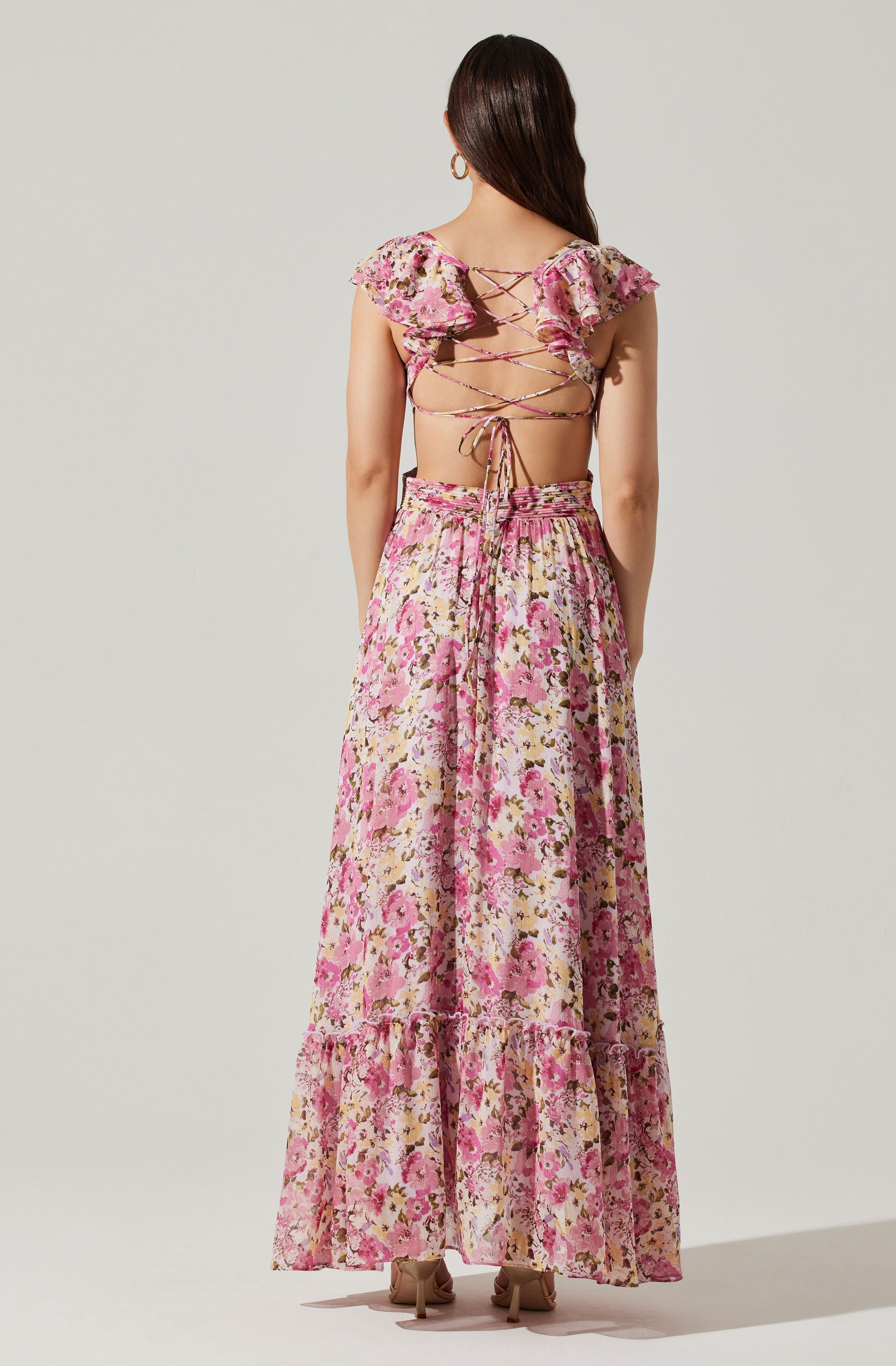 A woman wearing a pink floral two-piece outfit featuring a backless top with ruffled sleeves and crisscrossing strings, paired with a long, flowy skirt with a pink, beige, and green floral pattern.