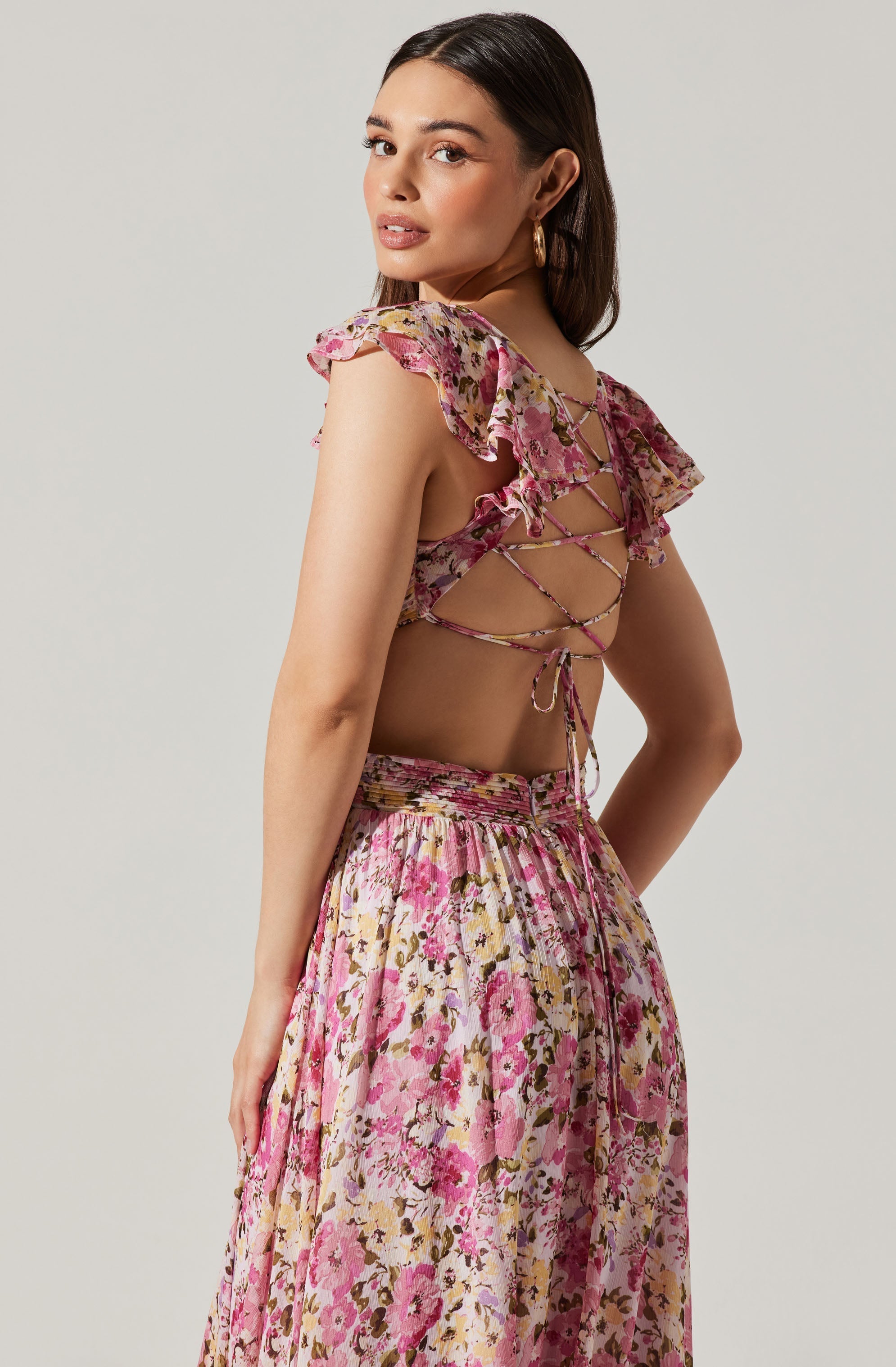 A woman is wearing a floral two-piece outfit with a ruffled, off-the-shoulder crop top that has a lace-up back. The top features pink, beige, and brown floral patterns with ruffled sleeves. The matching skirt has a high waist, is flowing, and is patterned with the same pink, beige, and brown floral design.