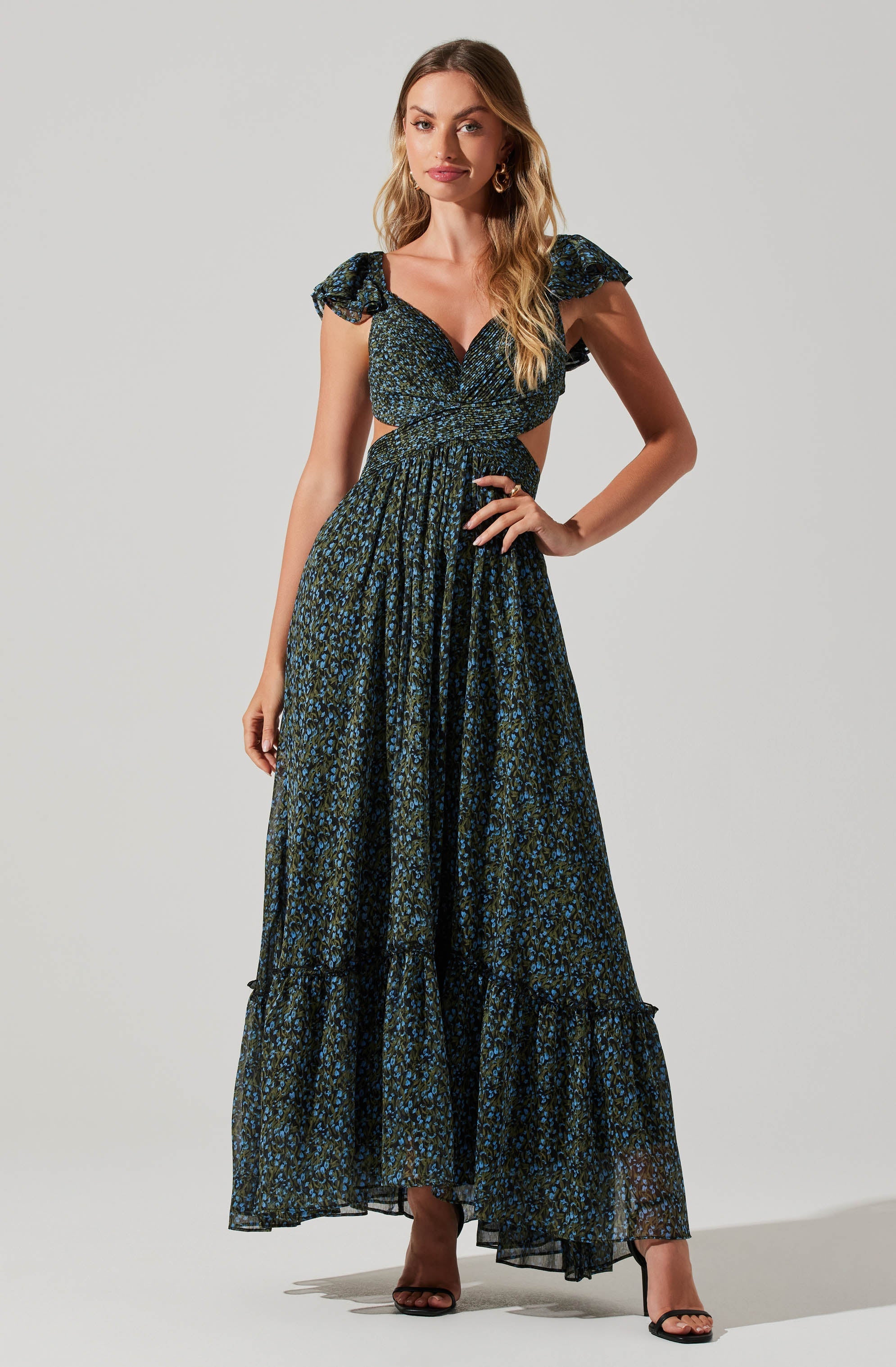 A woman is wearing a long, flowing dress with a dark base color and a blue and green floral pattern. The dress features ruffled short sleeves, a fitted bodice with a cross-over design, and cut-out side details at the waist. The skirt is tiered and reaches the floor, paired with open-toe black high heels. She has wavy blonde hair and is accessorized with large, round earrings.
