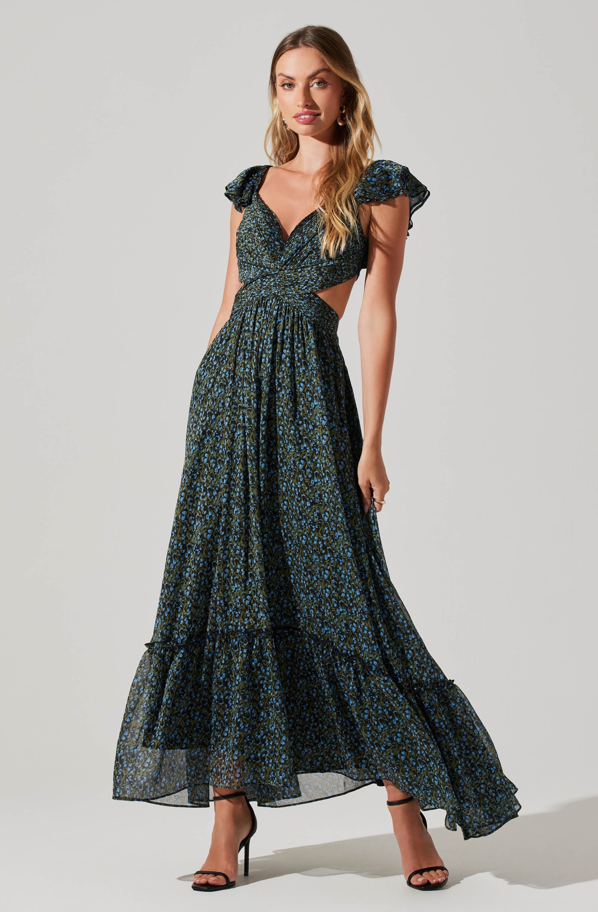 A woman is wearing a flowy, floor-length dress with a dark blue base color and a small, scattered blue and green floral pattern. The dress features ruffled cap sleeves, a sweetheart neckline, and cut-outs on the sides just below the bust. She pairs it with black high-heeled sandals.
