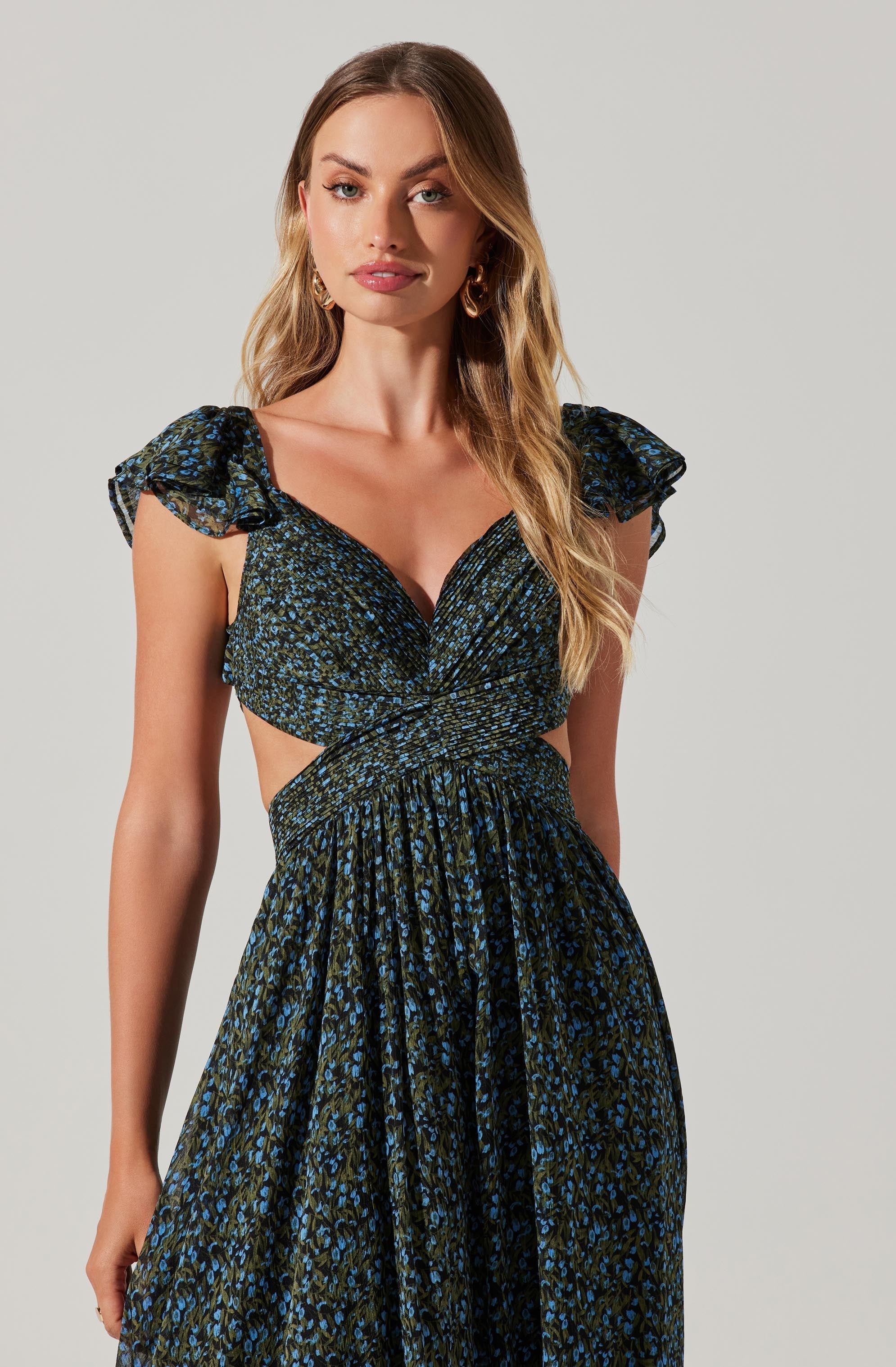 A woman wearing a dark-colored dress with a blue and green floral pattern. The dress features ruffled, short sleeves, a wrap-style top with a deep neckline, and cutouts on the sides, exposing her midriff. The skirt is flowy and gathered at the waist.