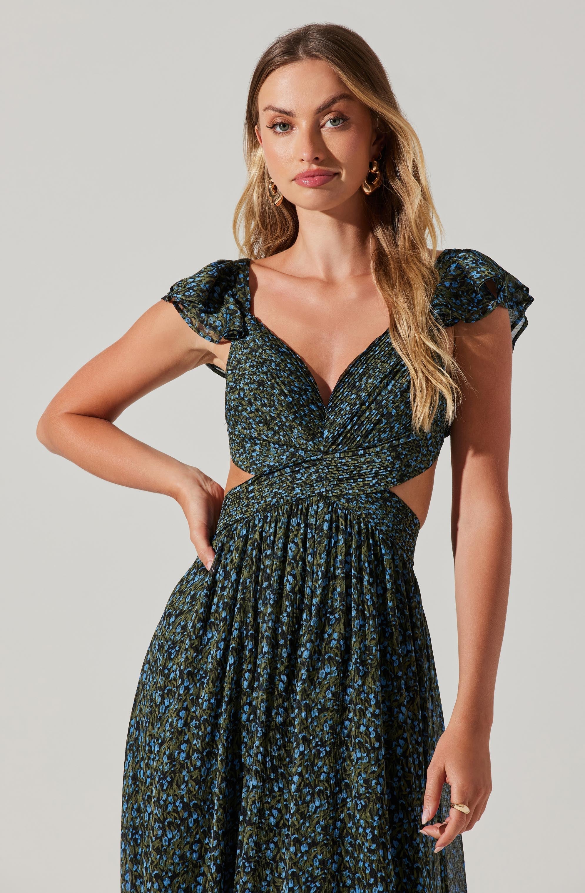 A woman is wearing a black dress with a blue and green floral pattern. The dress features short, ruffled sleeves, a deep V neckline, a cutout at the waist, and a high-waisted, gathered skirt.