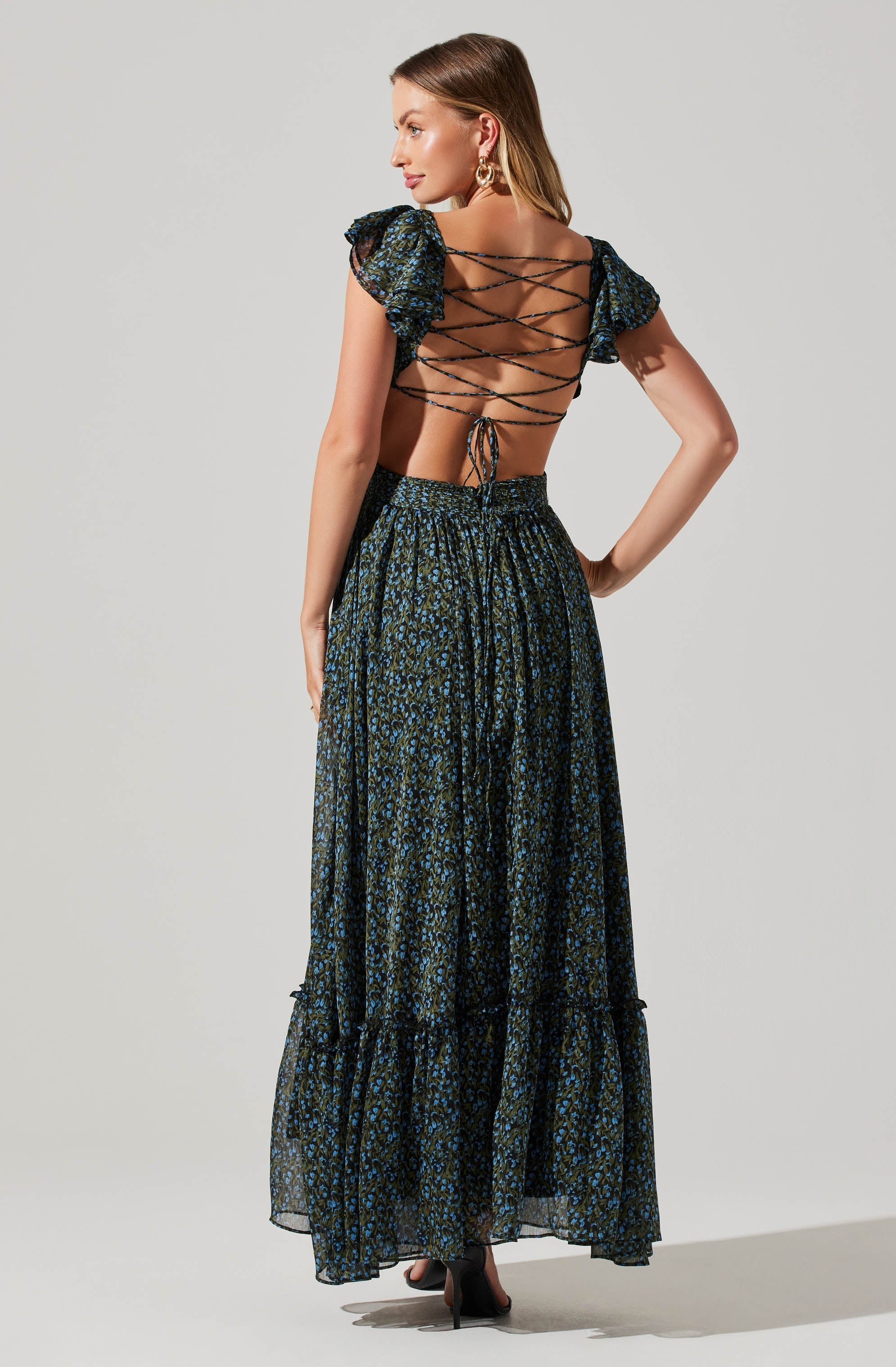 A woman is wearing a stylish two-piece outfit with a dark, floral-patterned fabric. The top features short puffed sleeves and a unique back design with crisscrossed laces, leaving much of her back exposed. The matching high-waisted skirt is long and flowing, with a similar floral pattern and a slightly ruffled hem. She is also wearing black high heels and gold hoop earrings.