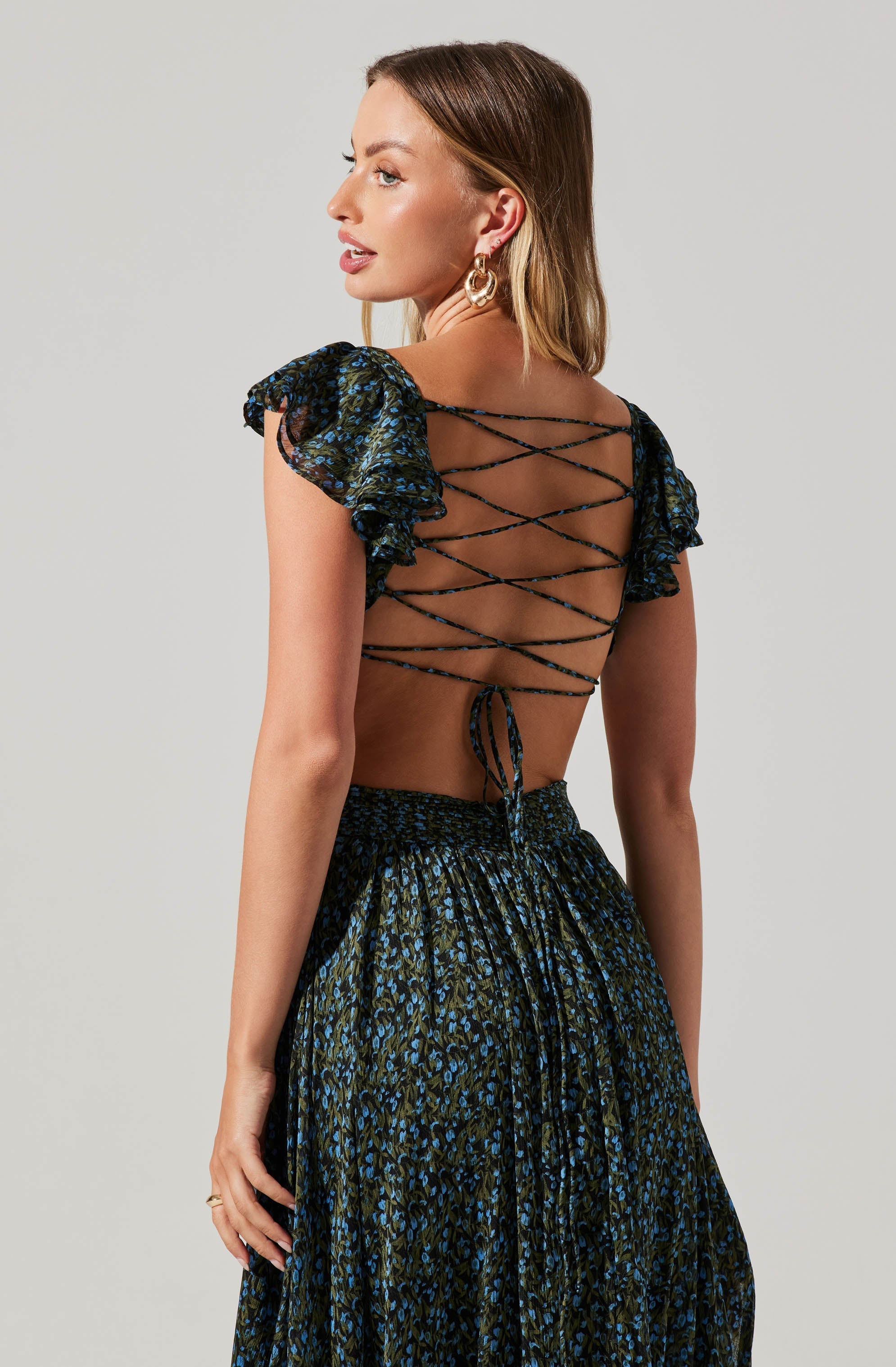 A woman is wearing a dark-colored dress with a floral pattern in blue and green. The dress features ruffled, puffed sleeves and a back design with crisscrossing laces that tie at the bottom.