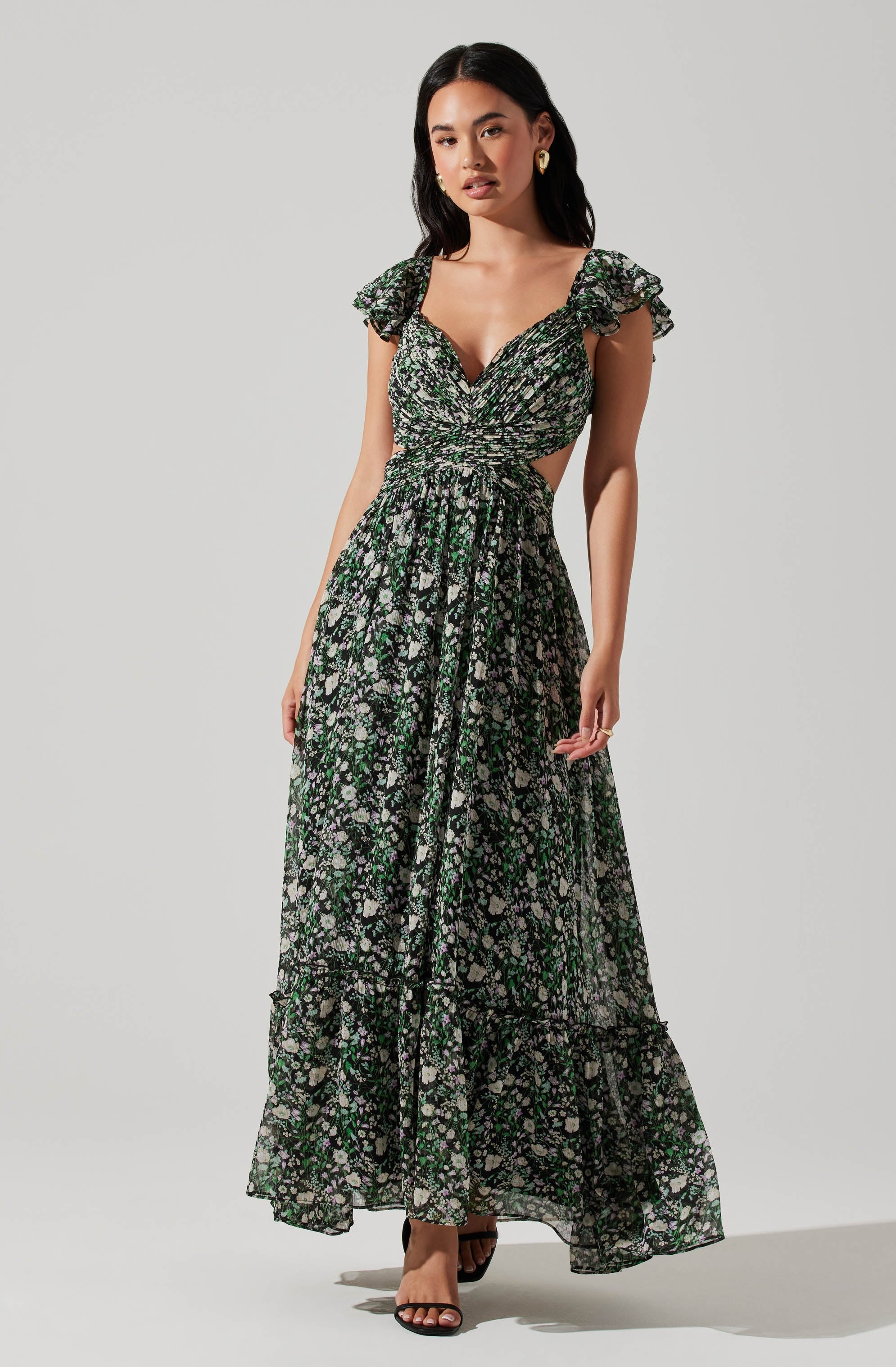 A woman is wearing a long, flowy maxi dress with a dark background and a small floral print in white, light purple, and green. The dress features ruffled cap sleeves, a gathered bodice with a sweetheart neckline, and cut-out sides.