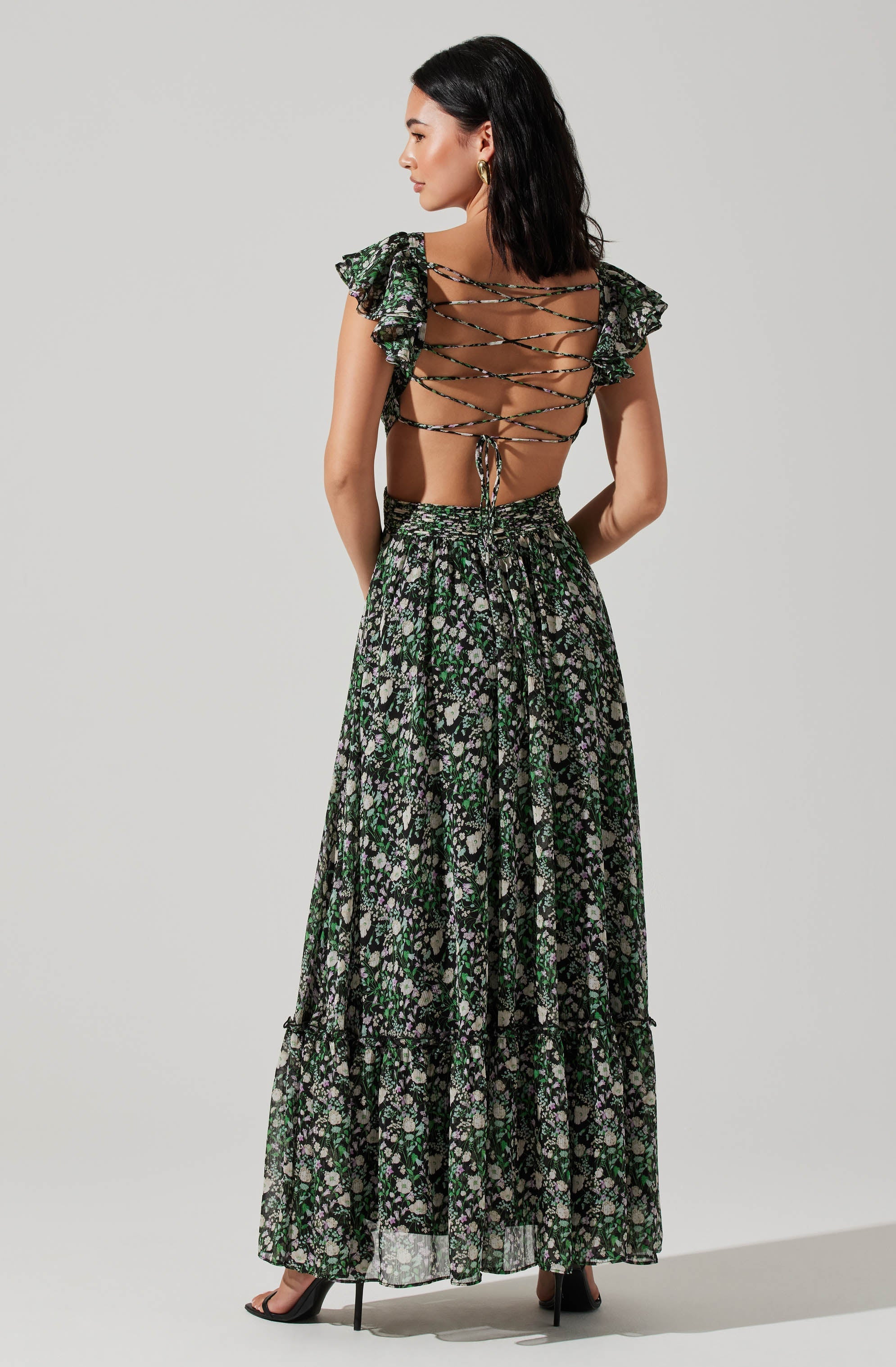 A woman is wearing a black floral maxi skirt with green and purple accents, paired with a backless top that features ruffled cap sleeves and crisscross laces up the back. She is also wearing black high-heeled shoes.