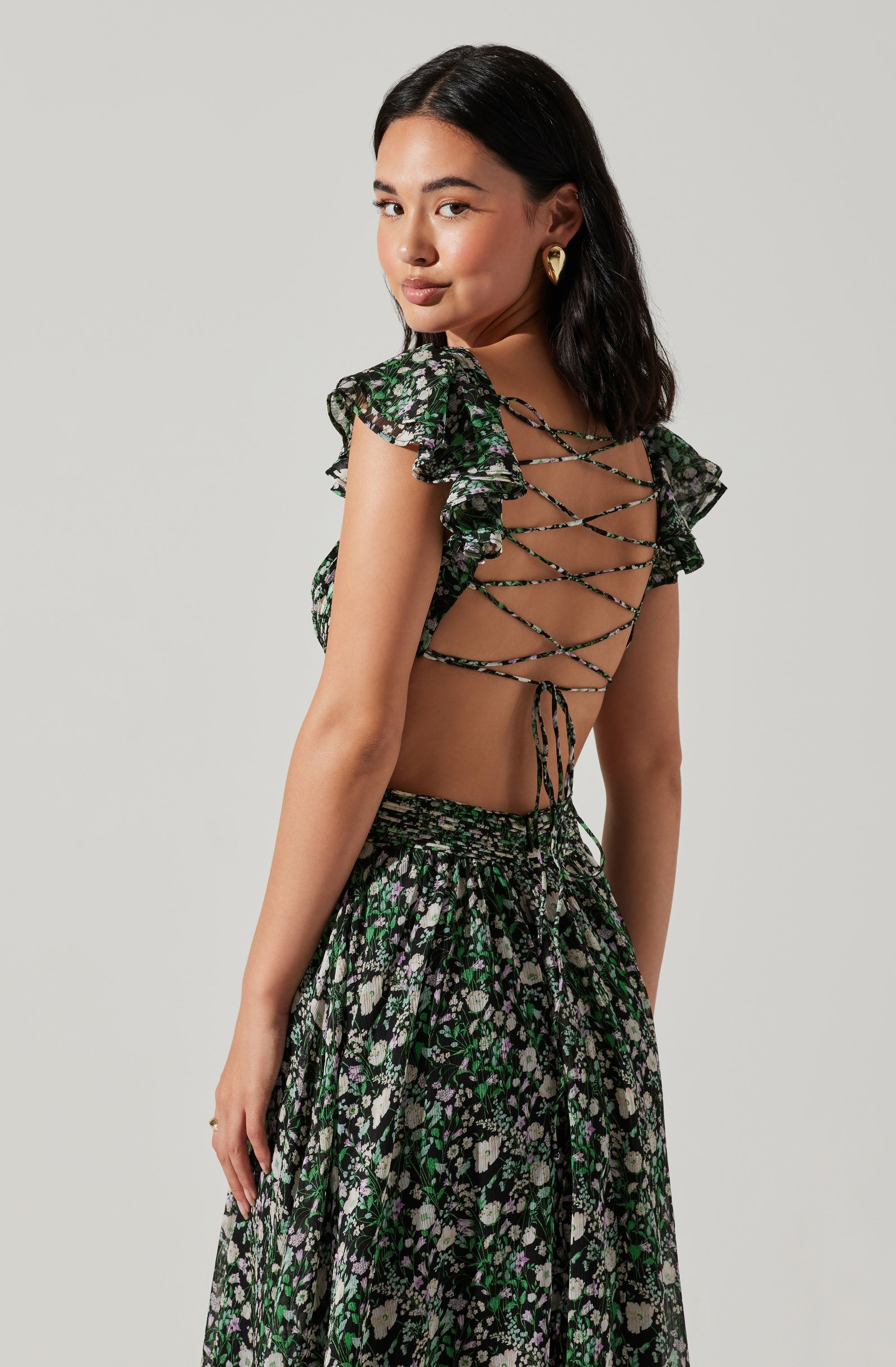 A young woman is wearing a black floral two-piece outfit. The top features short ruffled sleeves and a lace-up back with crisscrossing strings. The matching skirt is high-waisted with a loose, flowing fit, and is decorated with a small floral pattern in shades of green, purple, and beige. She is also wearing gold earrings and has shoulder-length black hair.