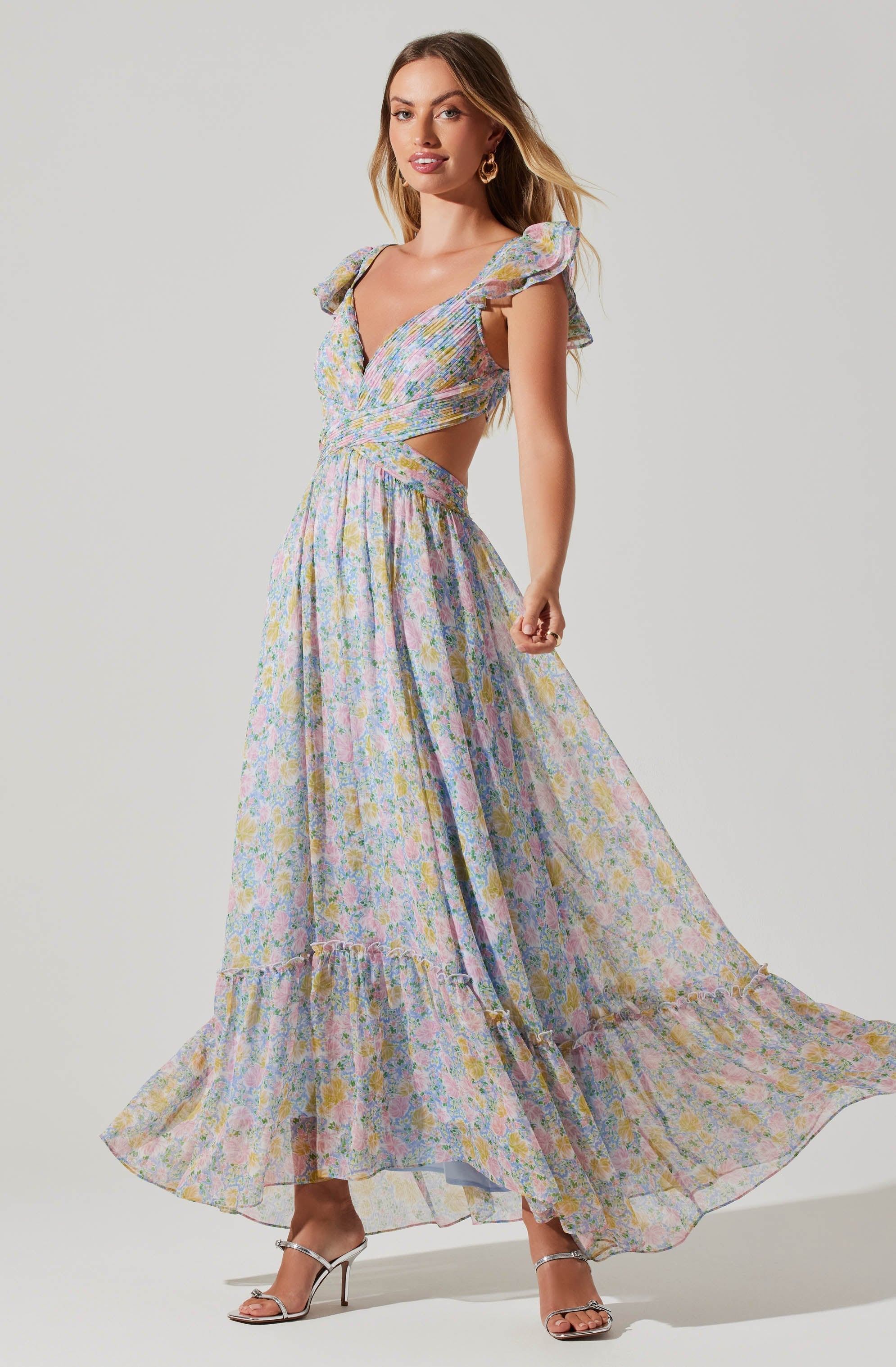 A woman is wearing a long, flowy dress with a pastel floral pattern. The dress features ruffled cap sleeves, a deep V neckline, and cut-out sides at the waist. The skirt is full-length with a gathered ruffle at the hem. She is also wearing metallic high-heeled sandals and gold hoop earrings.