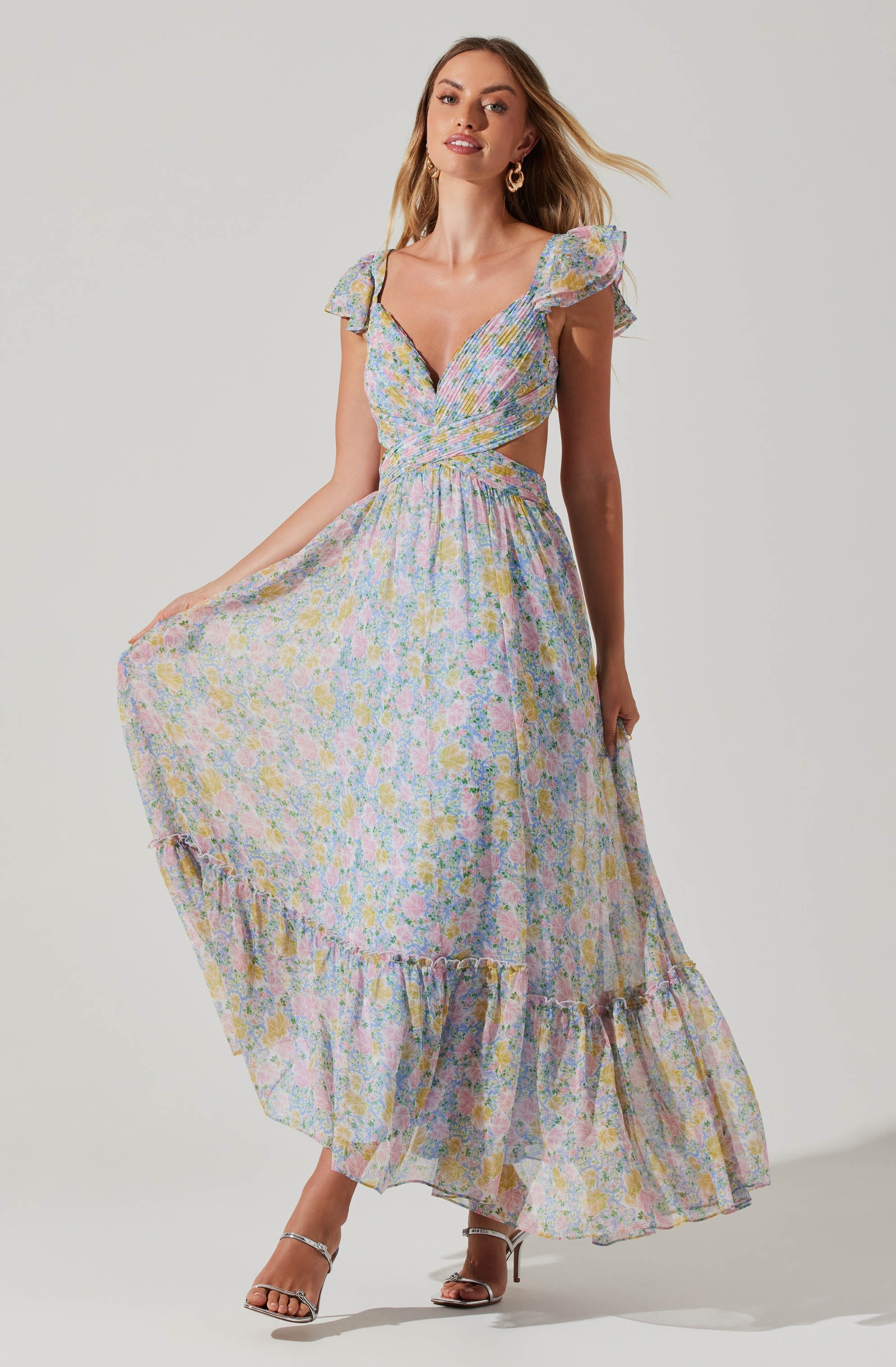 A woman is wearing a long, flowy dress with a colorful floral pattern. The dress has a deep V neckline, ruffled cap sleeves, and cutouts at the waist. The skirt features a ruffled hemline that reaches her ankles. She is paired with strappy high-heeled sandals.