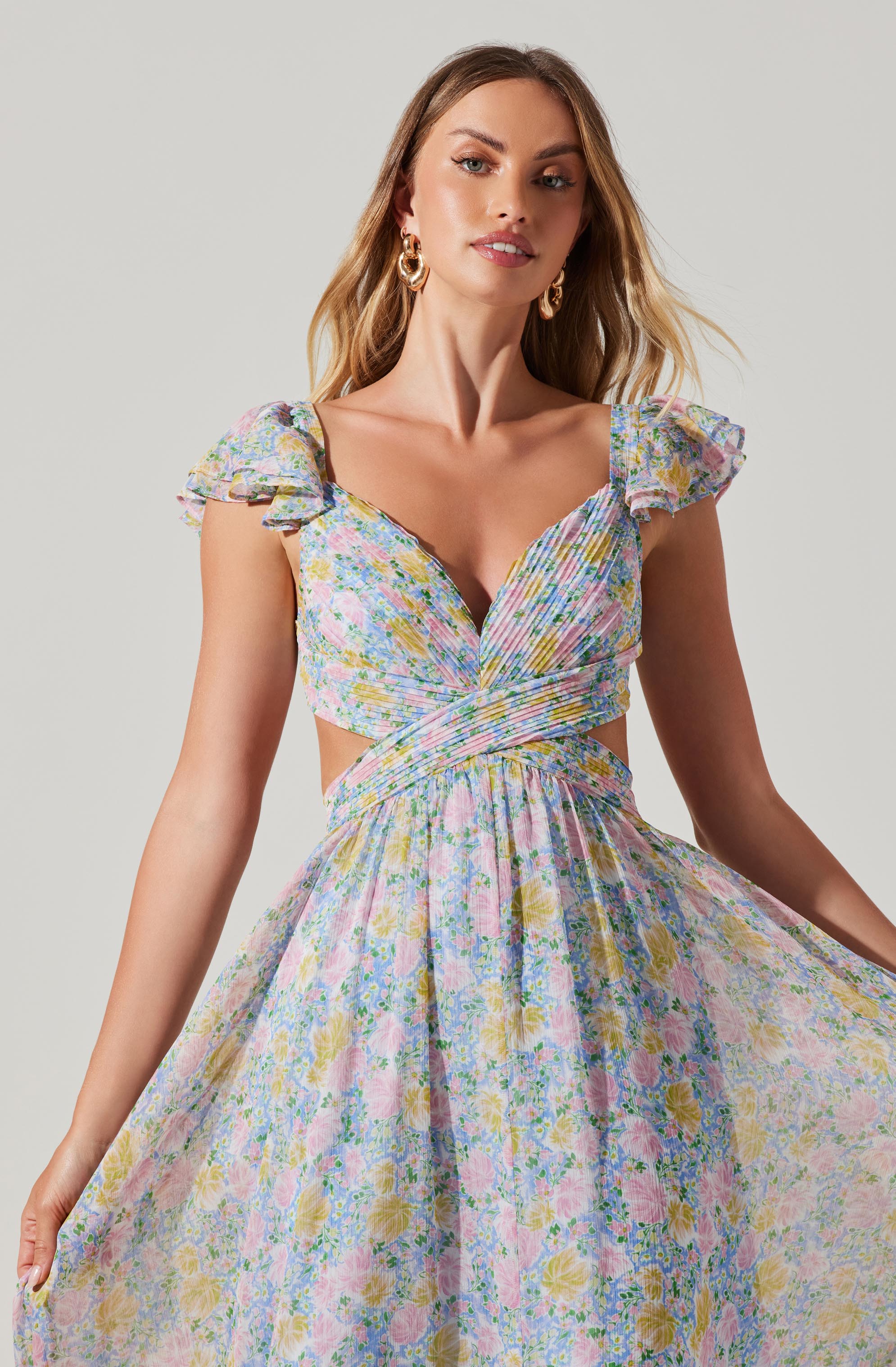 A woman is wearing a light, pastel-colored dress with a floral pattern. The dress features a fitted bodice with a deep neckline and crisscrossing fabric at the waist, creating a cutout effect at the sides. The dress has short, ruffled sleeves and a flowing, gathered skirt. She accessorizes with large, gold hoop earrings.