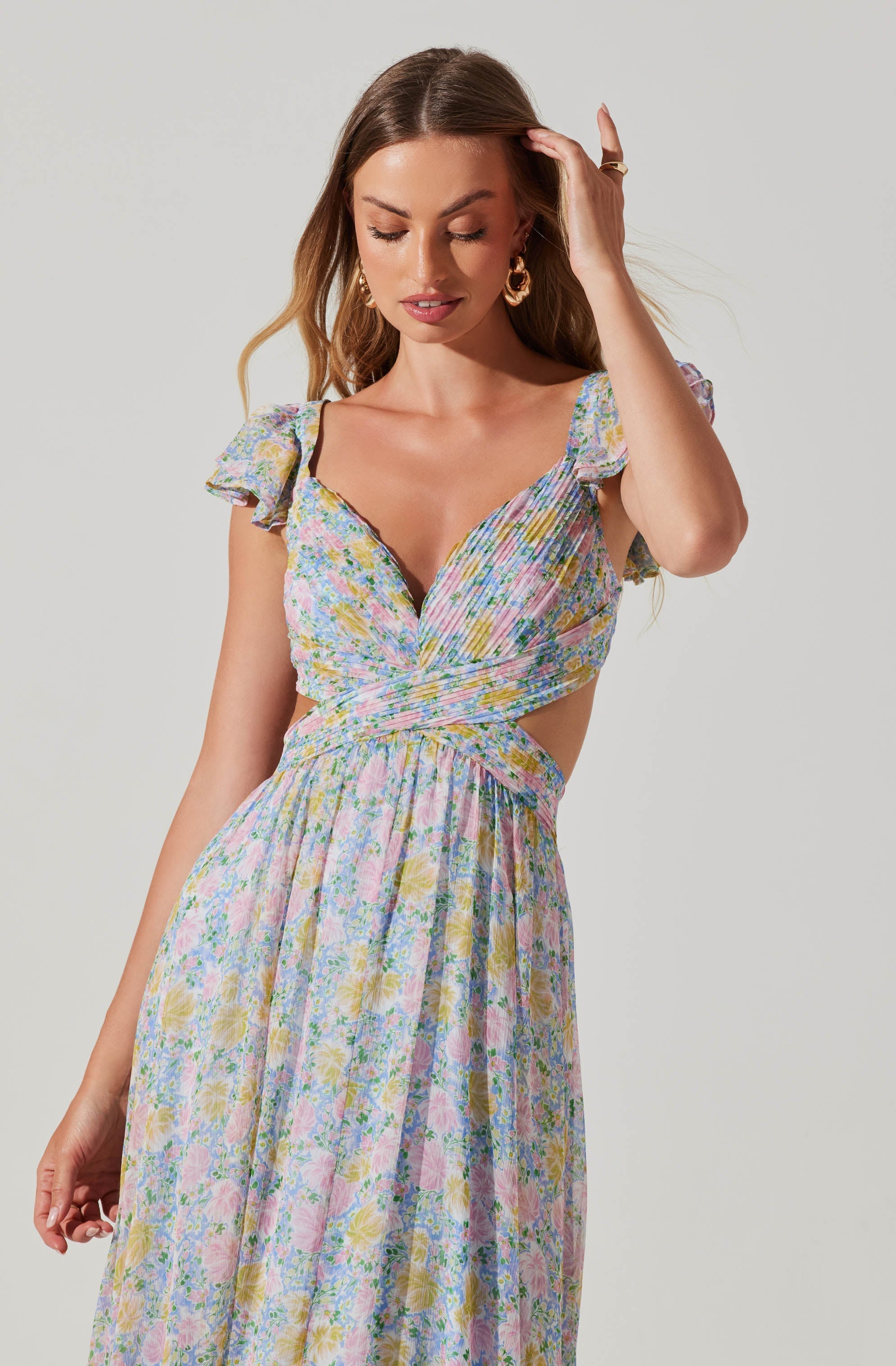 A woman wearing a colorful floral dress with a smocked waistband. The dress has ruffled short sleeves, a deep V neckline, and a cutout at the midriff. The fabric features a pastel floral pattern with shades of pink, yellow, blue, and green.