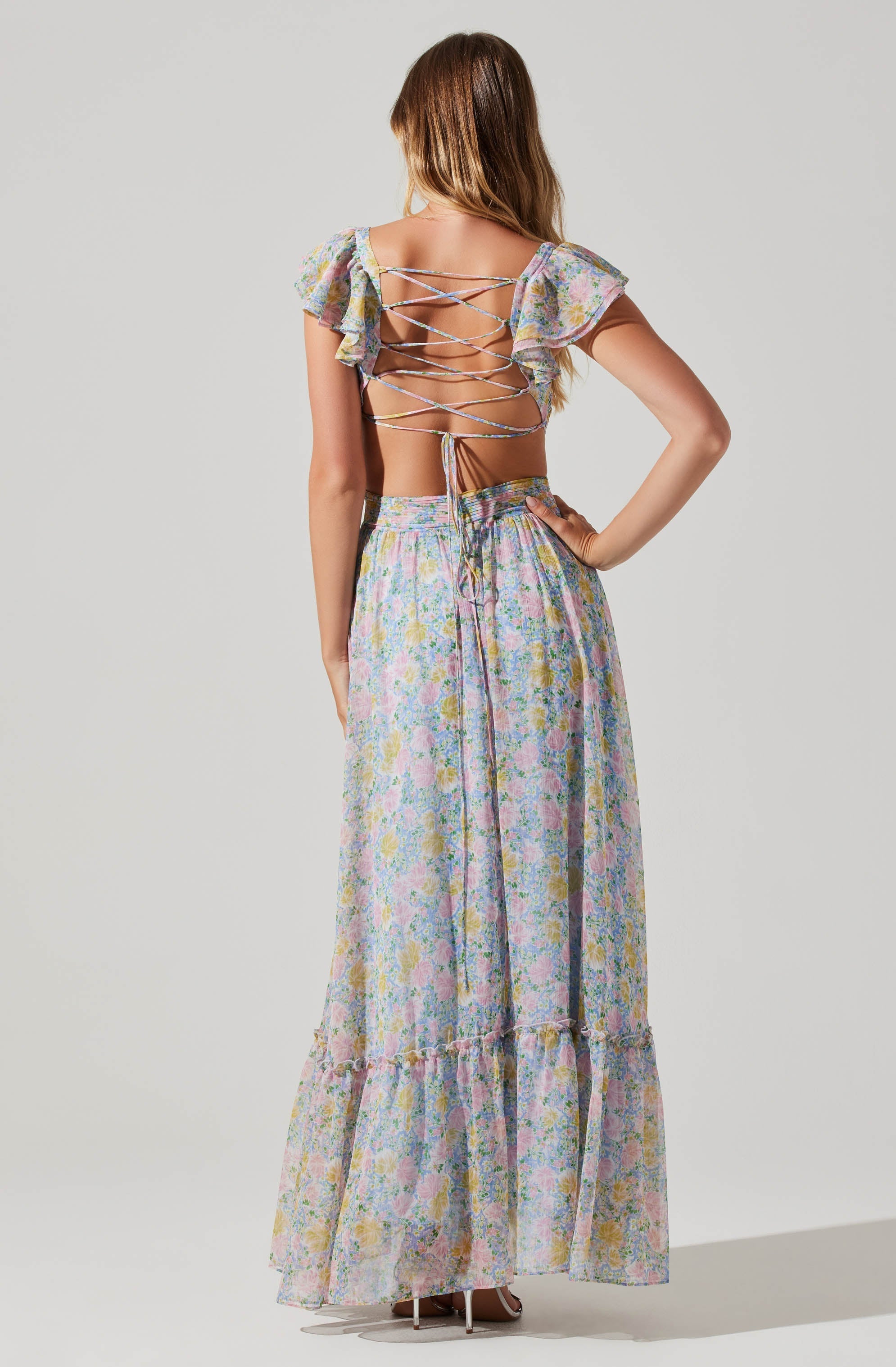 A woman is wearing a two-piece outfit with a floral pattern in pastel colors. The top has ruffled sleeves and a corset-style back with crisscrossing strings. The bottom is a long, flowy skirt with a ruffled hem.