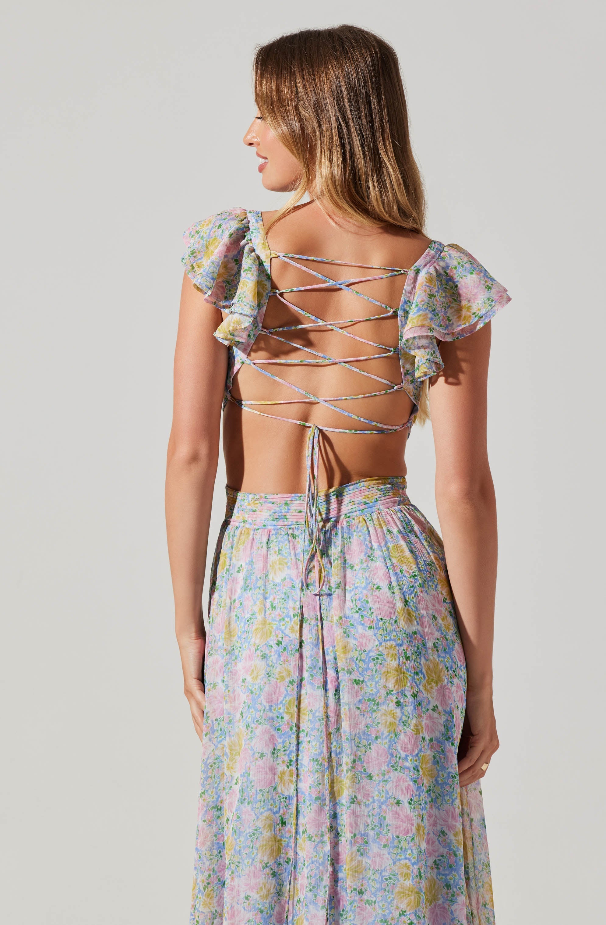 A woman is wearing a backless top with thin, crisscrossing straps and short, ruffled sleeves made of a sheer, floral-patterned fabric. She pairs it with a high-waisted, flowing skirt featuring a colorful floral print.