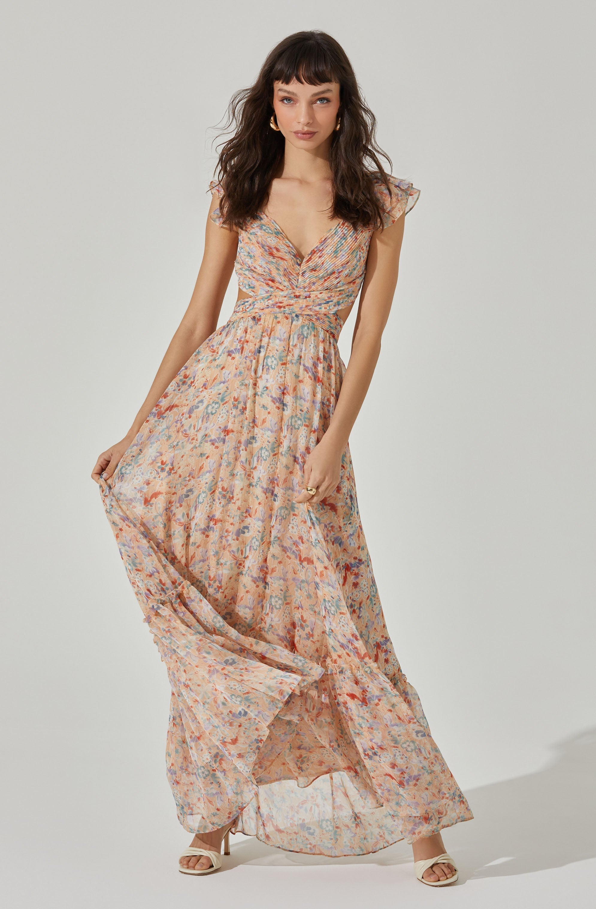 A woman is wearing a long, flowy dress with a floral pattern in shades of peach, blue, and purple. The dress has a V-neckline, short flutter sleeves, and a fitted waist that accentuates her figure. She is also wearing white open-toe heels with ankle straps.