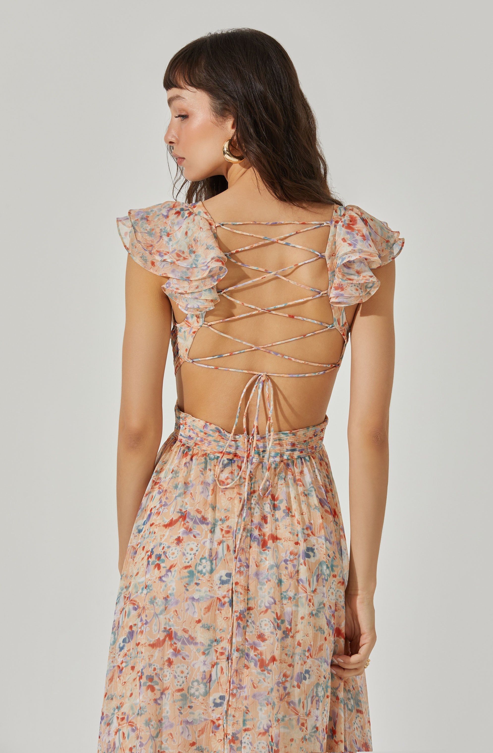 A woman is wearing a backless dress with short, ruffled sleeves and a floral pattern in soft pastel colors. The dress features crisscrossing laces on the back that tie at the bottom, revealing her back and shoulders. The skirt of the dress is high-waisted and flows down, matching the floral pattern and colors of the top.