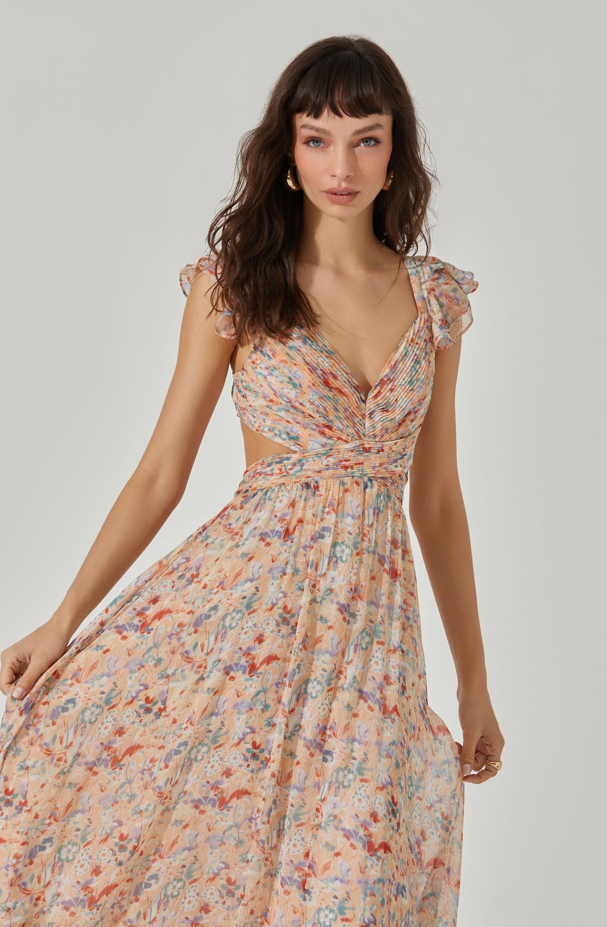 A woman is wearing a flowy, floral-patterned dress in shades of peach, blue, and red. The dress features ruffled shoulder details, a deep V neckline, and a smocked waistline that accentuates her figure.