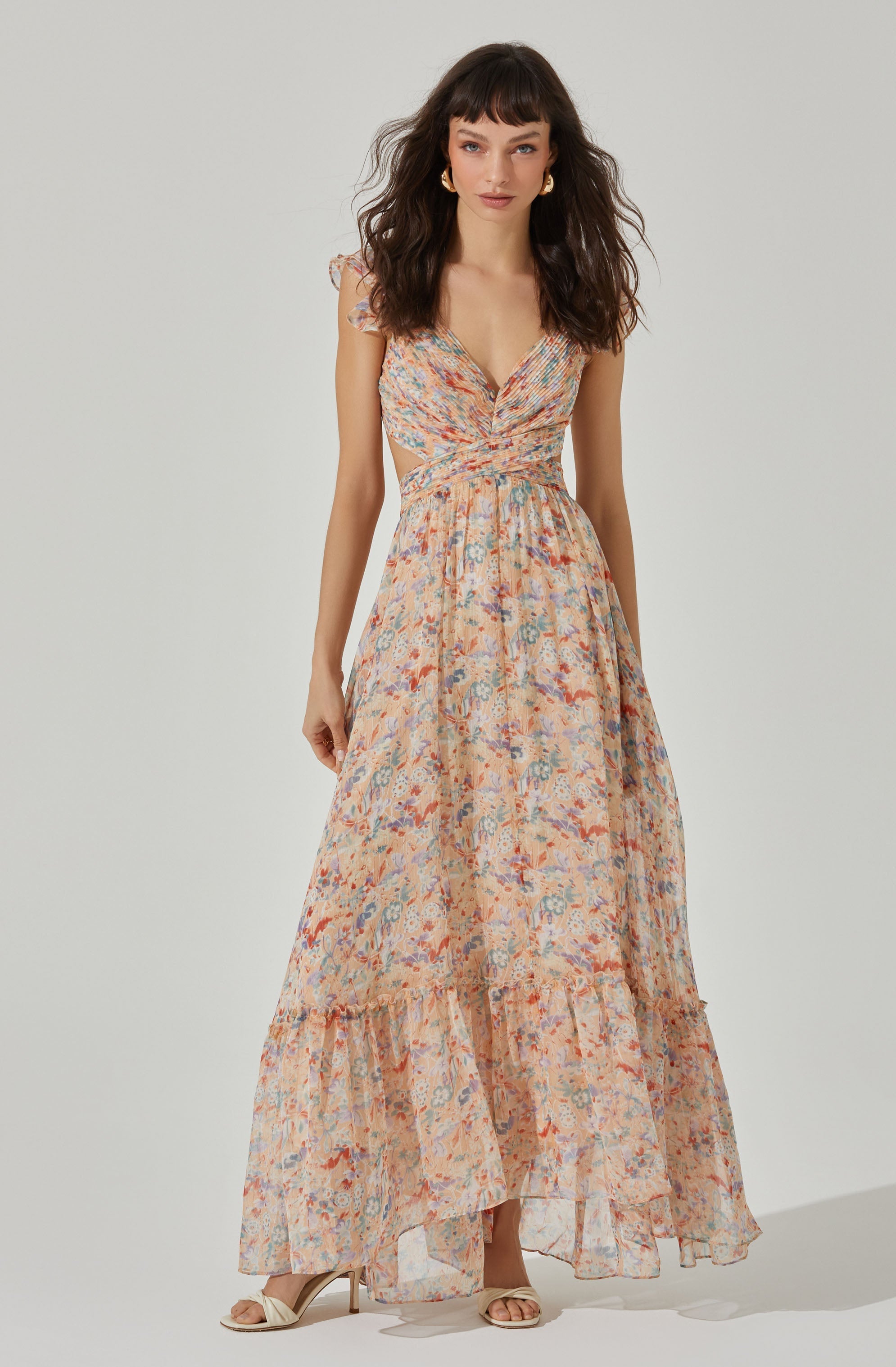 A woman is wearing a long, flowing dress with a colorful, floral pattern. The dress features a V-neckline, ruched detailing at the bodice, and ruffled shoulder straps. The skirt has tiered sections with ruffled trim near the hem. She is accessorized with gold hoop earrings and is wearing open-toe high-heeled shoes.