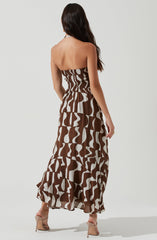 A woman is wearing a strapless, maxi dress with a fitted, ruched bodice. The dress features a bold, abstract pattern in shades of brown and beige, with a flowy, tiered skirt that reaches the floor. She is also wearing high-heeled sandals in a neutral tone.