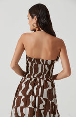 A woman is wearing a strapless dress with a brown and white geometric pattern. The dress has a smocked bodice that fits snugly around the top, and a gathered skirt that flows down from the waist. She has long, dark hair and is accessorized with large gold hoop earrings.