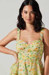 A woman is wearing a sleeveless, fitted dress with a yellow background and a multicolored floral print consisting of pink, green, orange, and purple. The dress has a square neckline with fabric-covered buttons running down the center. The fabric appears lightweight and has a gathered detail at the waist. She accessorized with large, gold, heart-shaped earrings and has dark, wavy, shoulder-length hair.