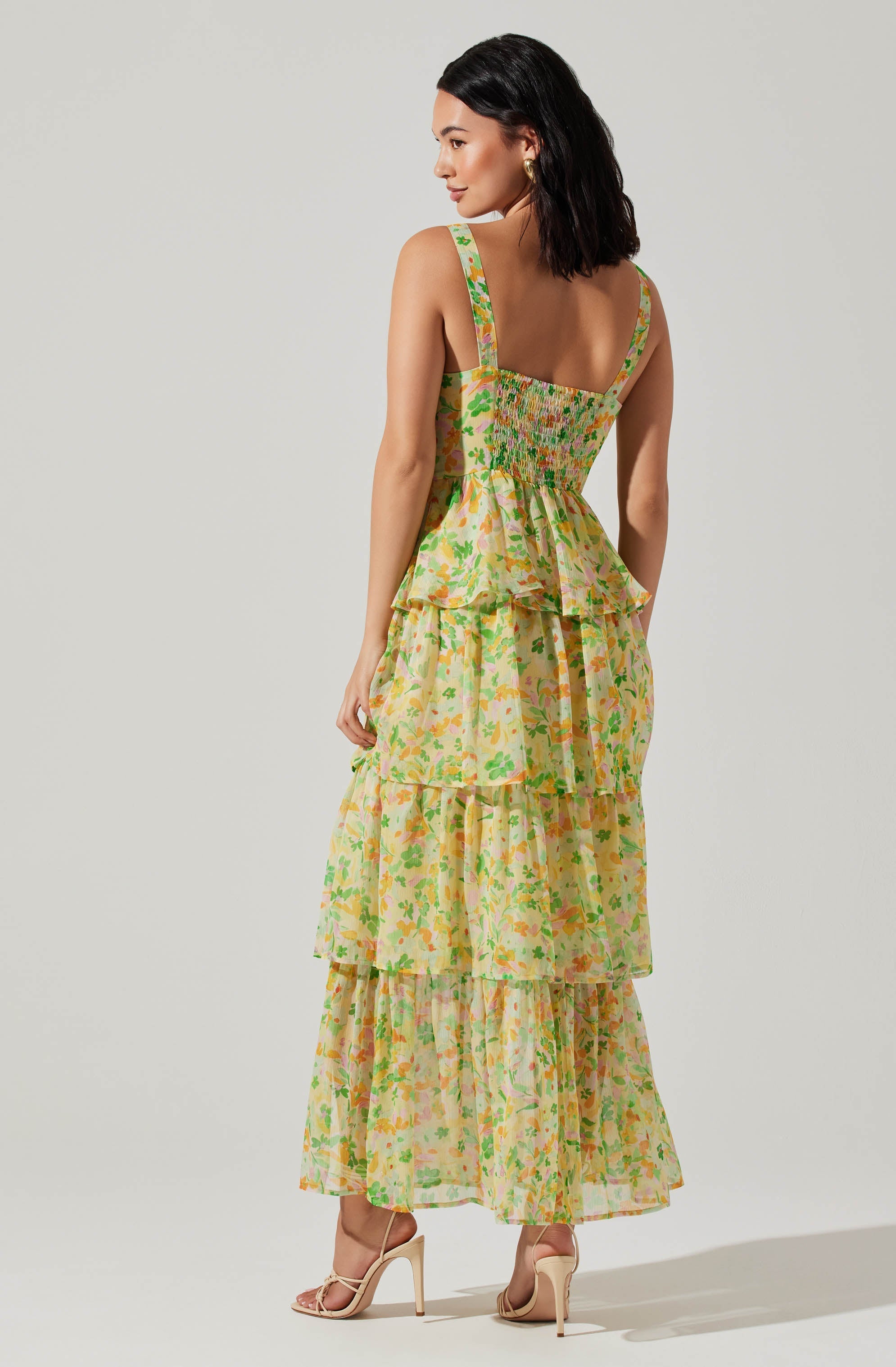 A woman is wearing a sleeveless, tiered maxi dress with a fitted, embroidered bodice. The dress features wide shoulder straps and a floral print with shades of yellow, green, pink, and orange. She is also wearing beige high-heeled sandals with ankle straps.
