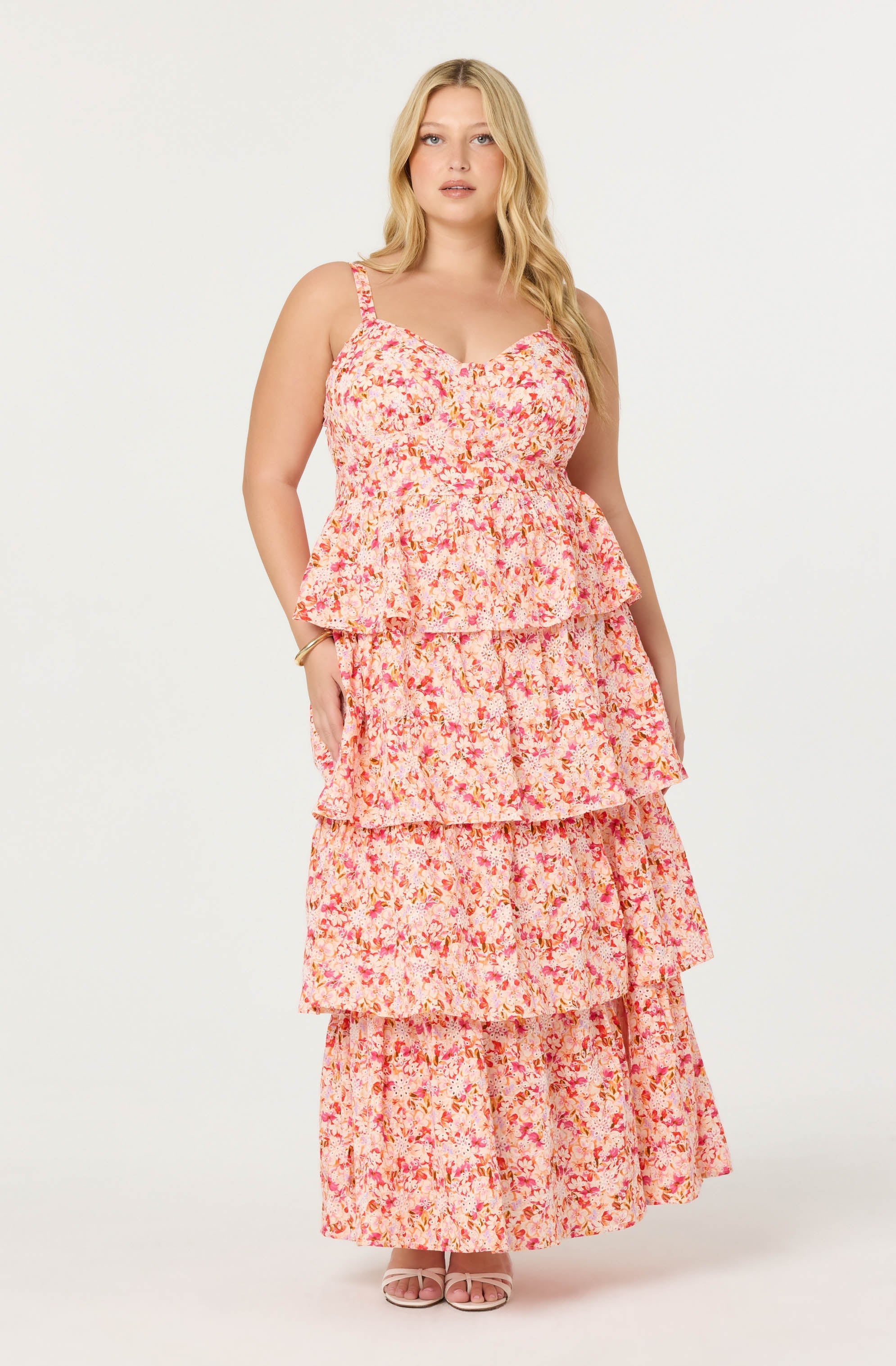 A woman is wearing a sleeveless, floral maxi dress with multiple tiered ruffles in shades of pink, red, and orange on a light background. The dress has thin spaghetti straps and a fitted bodice.