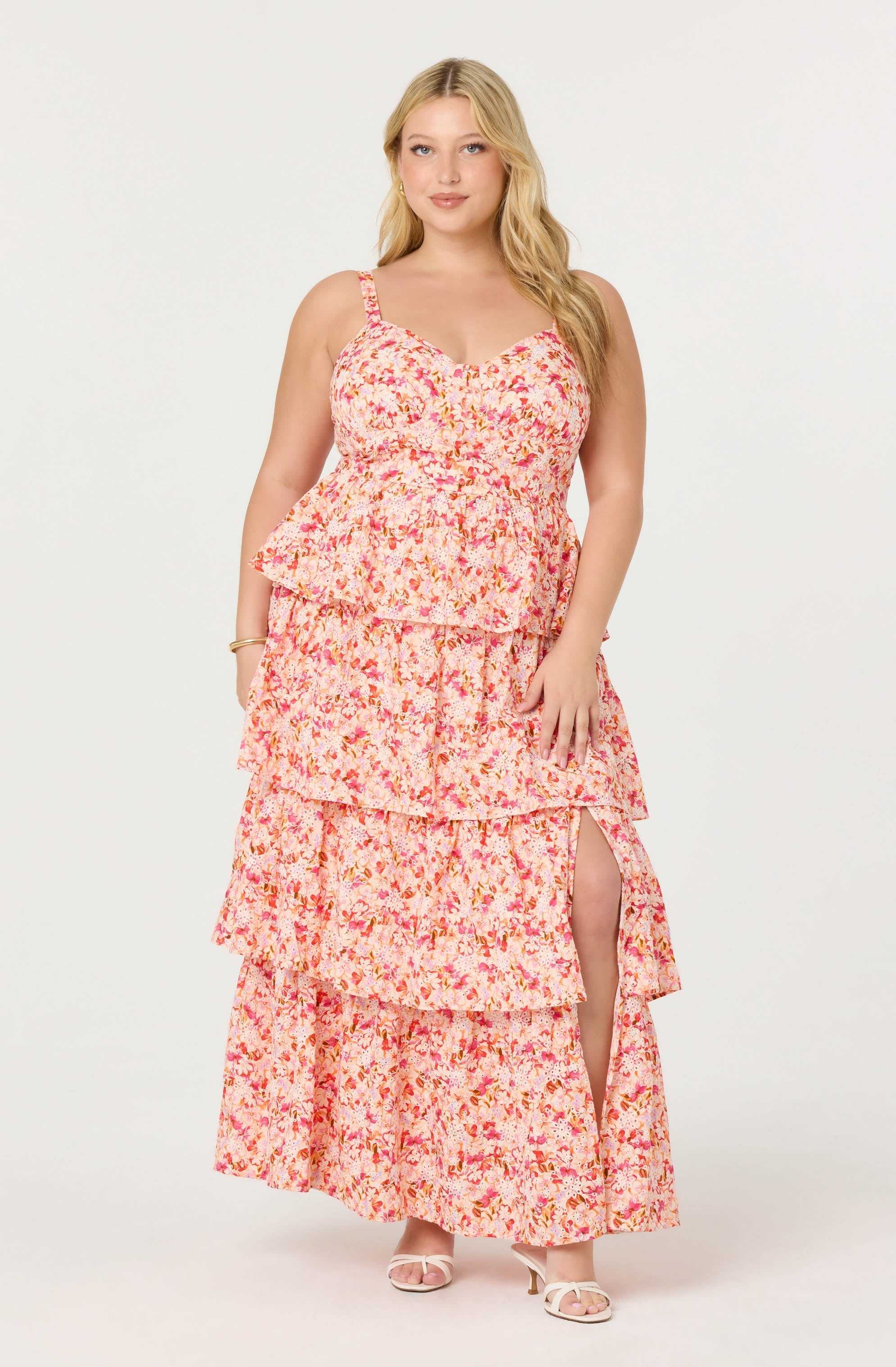 A woman is wearing a long, sleeveless, tiered maxi dress with a floral print featuring shades of pink, orange, and cream. The dress has a fitted bodice with thin shoulder straps, and a high slit on the right side revealing her leg. She is also wearing white open-toe heeled sandals.