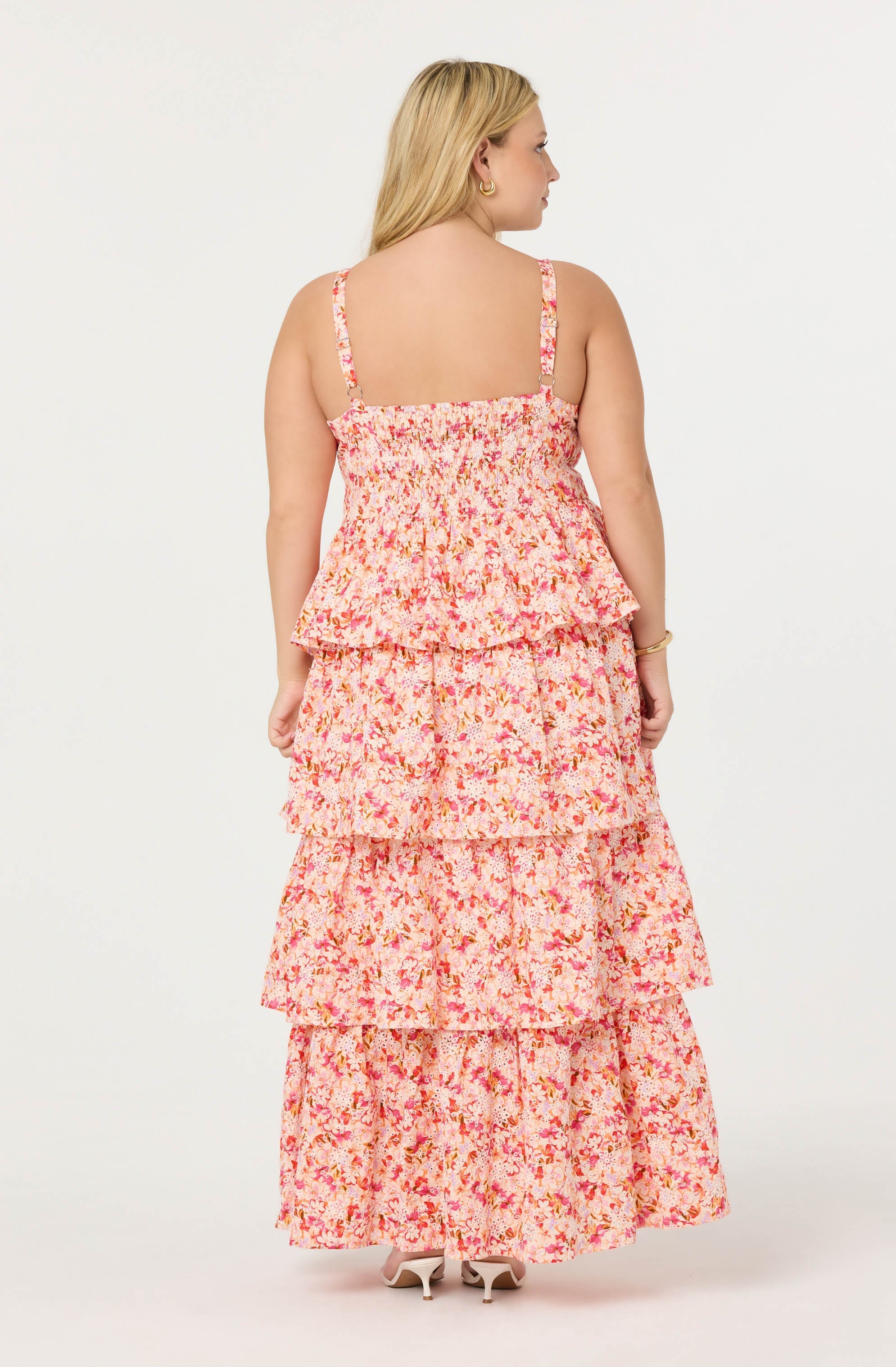 A woman is wearing a sleeveless, floral-print sundress with adjustable shoulder straps. The dress features a tiered, ruffled skirt in shades of pink, coral, and white. She is also wearing light-colored heels.